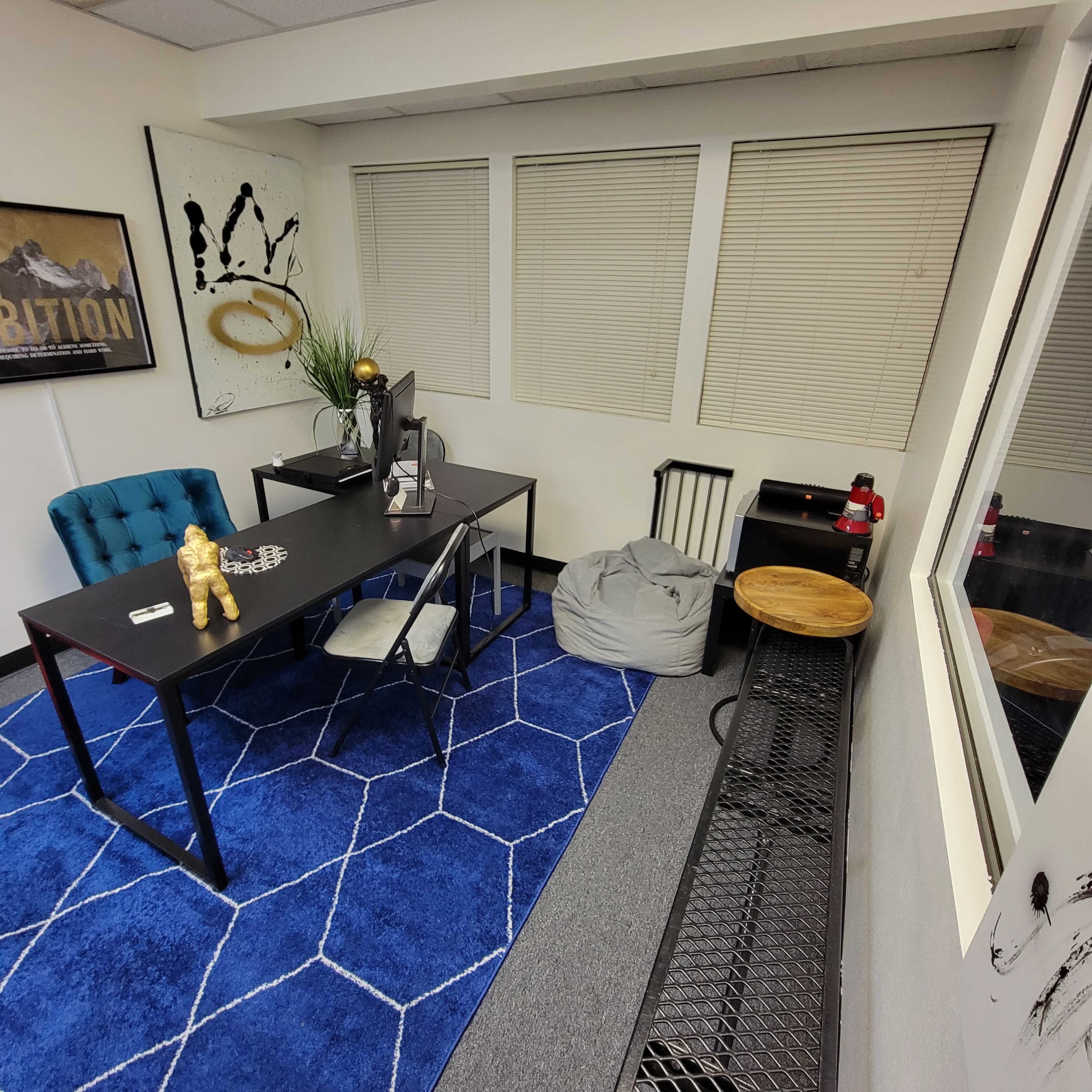 A small office space featuring a black desk, a blue rug with a geometric pattern, a bean bag chair, and a decorative wall art piece.