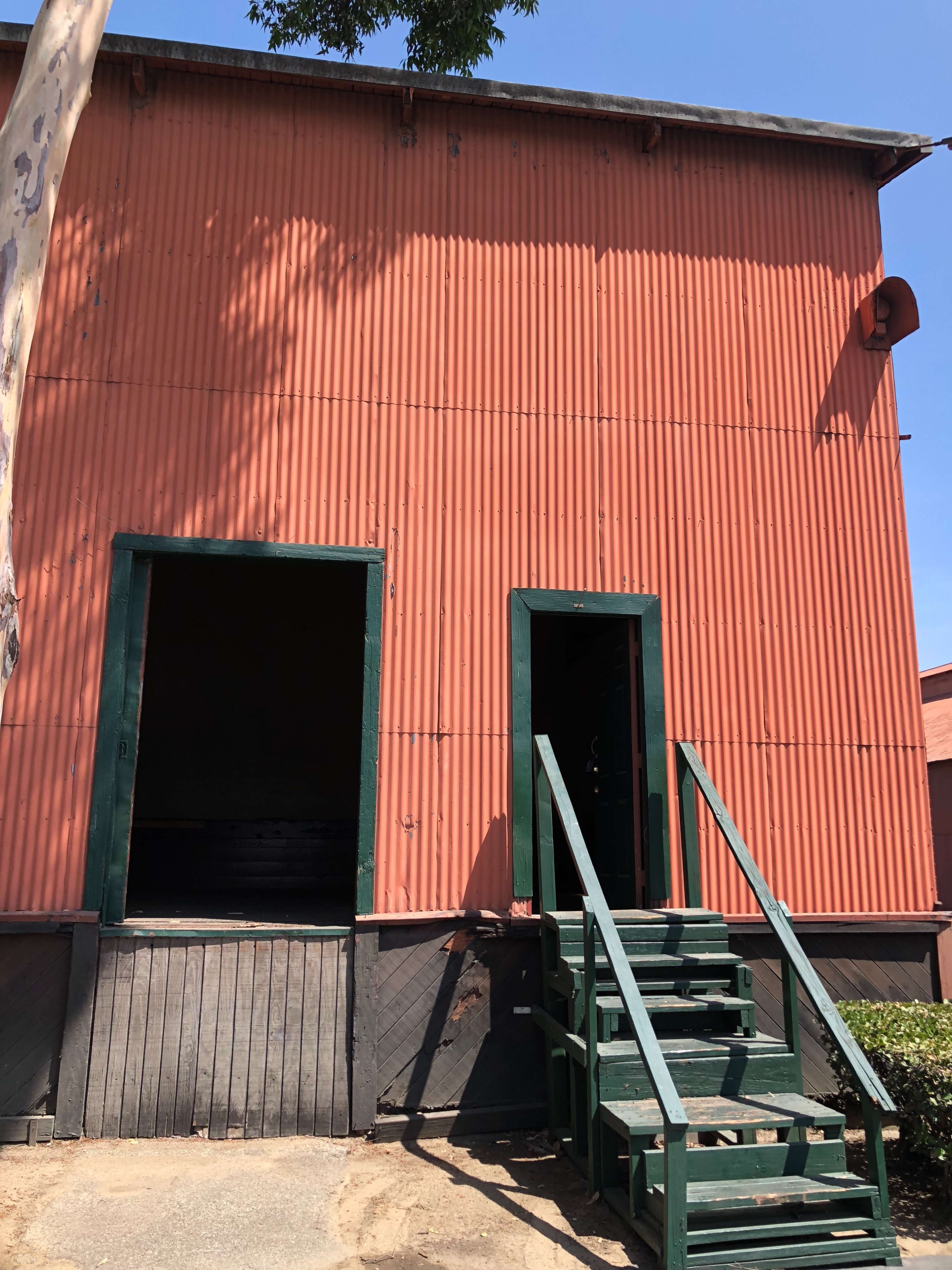 A red corrugated metal building with two open doors and a green staircase leading up to the entrance.