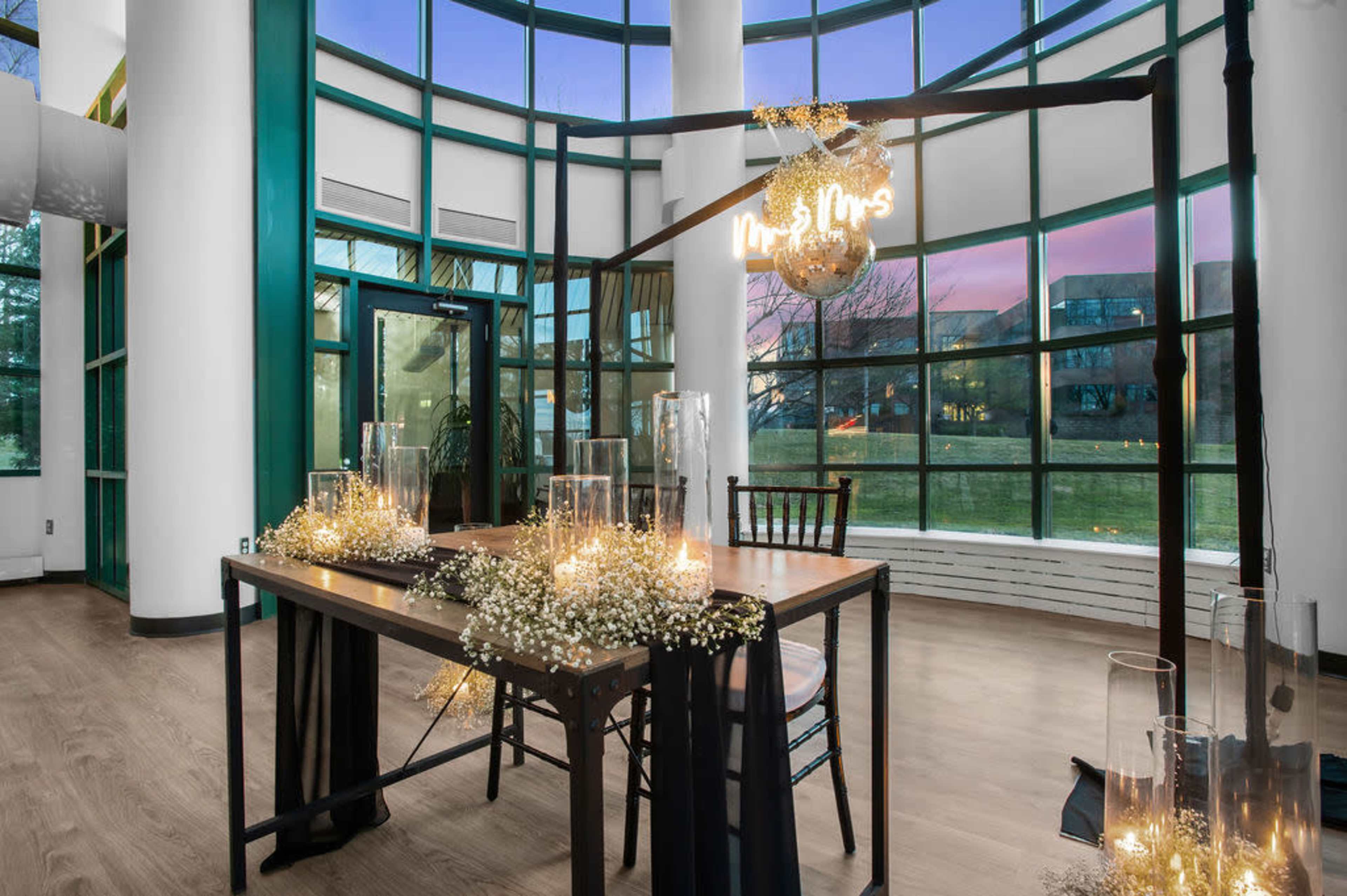 A decorated table with a "Mr. & Mrs." neon sign sits in a brightly lit room featuring large windows and a wooden floor.