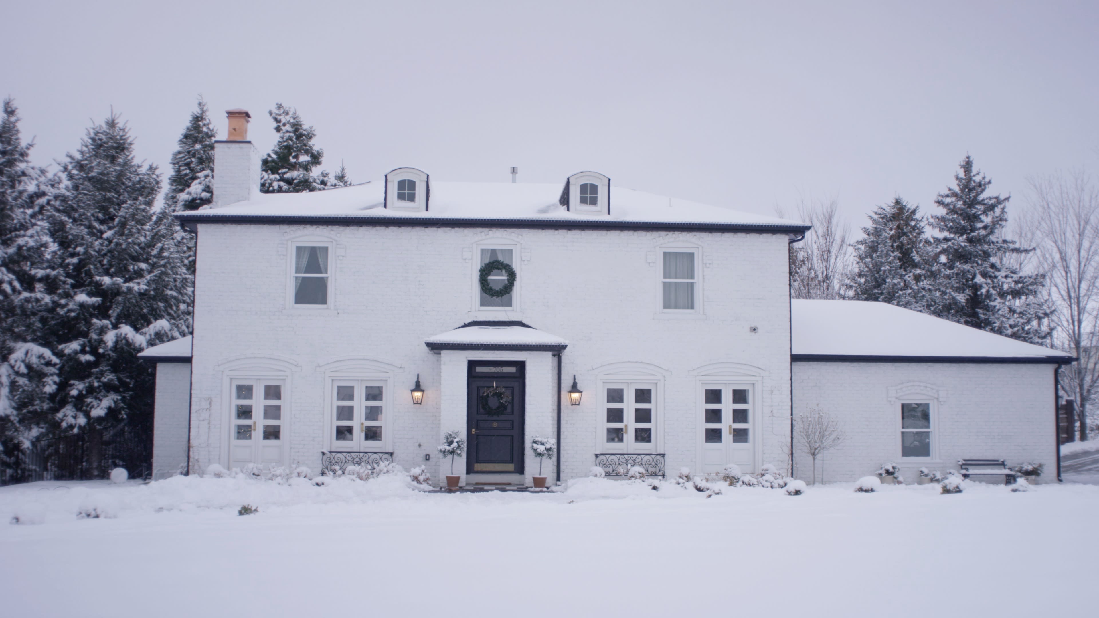 A two-story white house with dark trim is surrounded by snow-covered ground and trees, featuring a wreath on the front door and lights on either side.