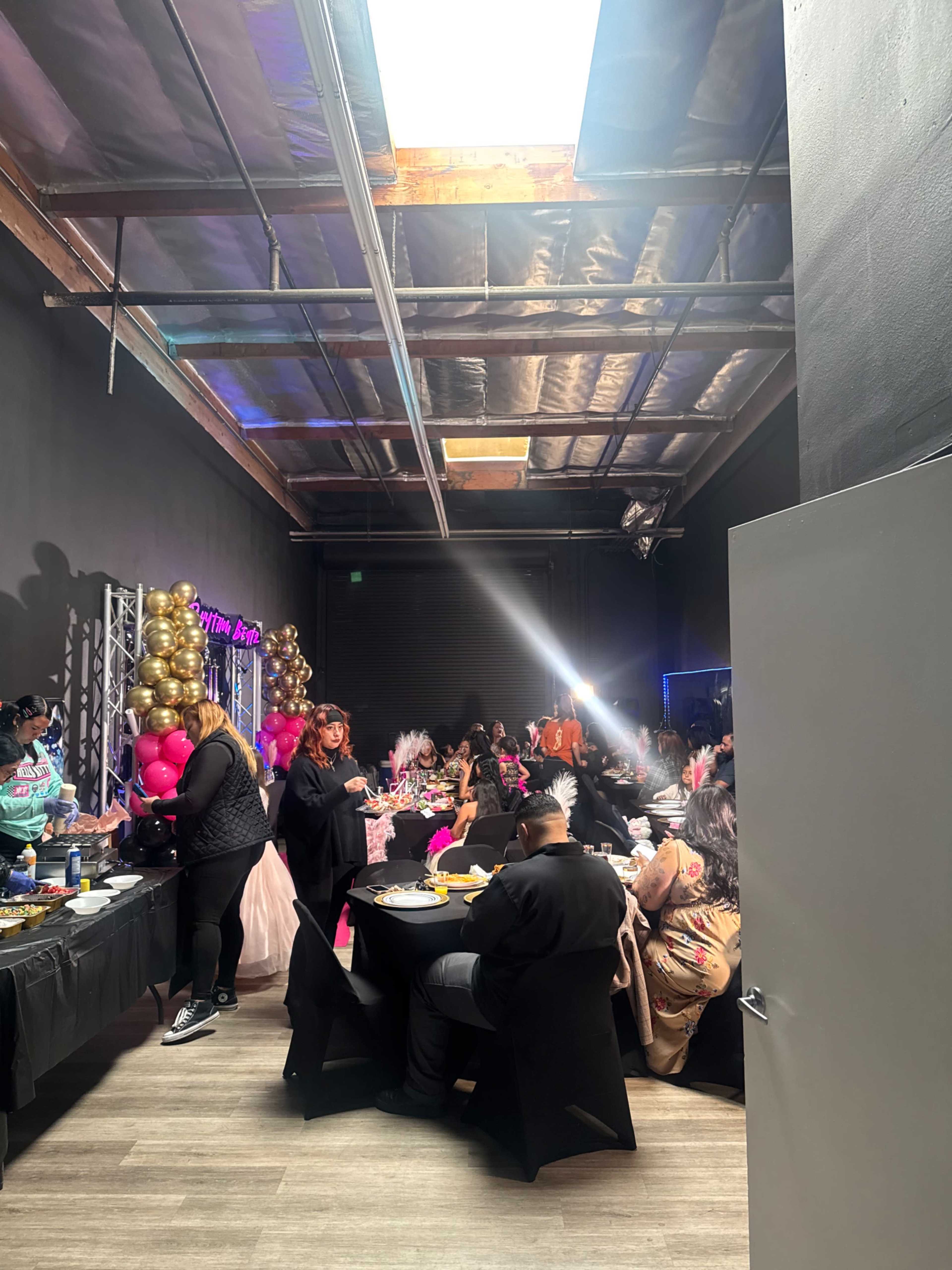 The image shows a lively indoor event with guests seated at tables, enjoying food and celebrating, surrounded by colorful decorations and balloons.