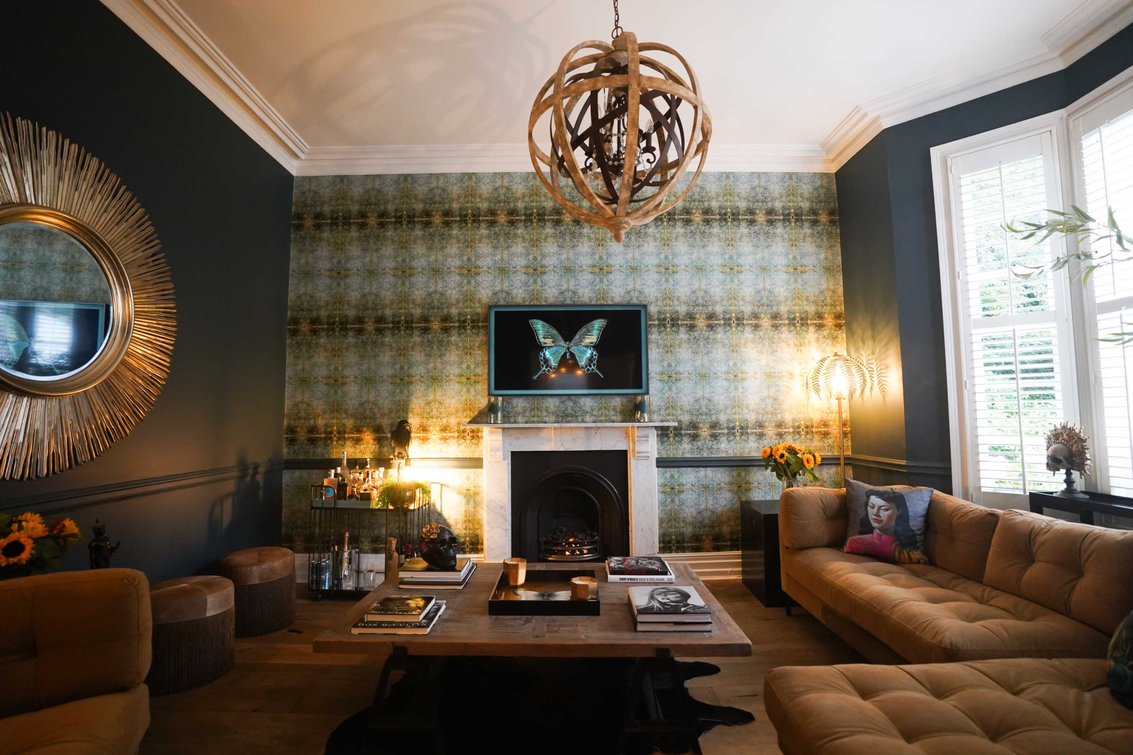The living room features a central coffee table, a large flat-screen TV mounted above a decorative fireplace, and brown sofas arranged around the space with patterned wallpaper and decorative elements.