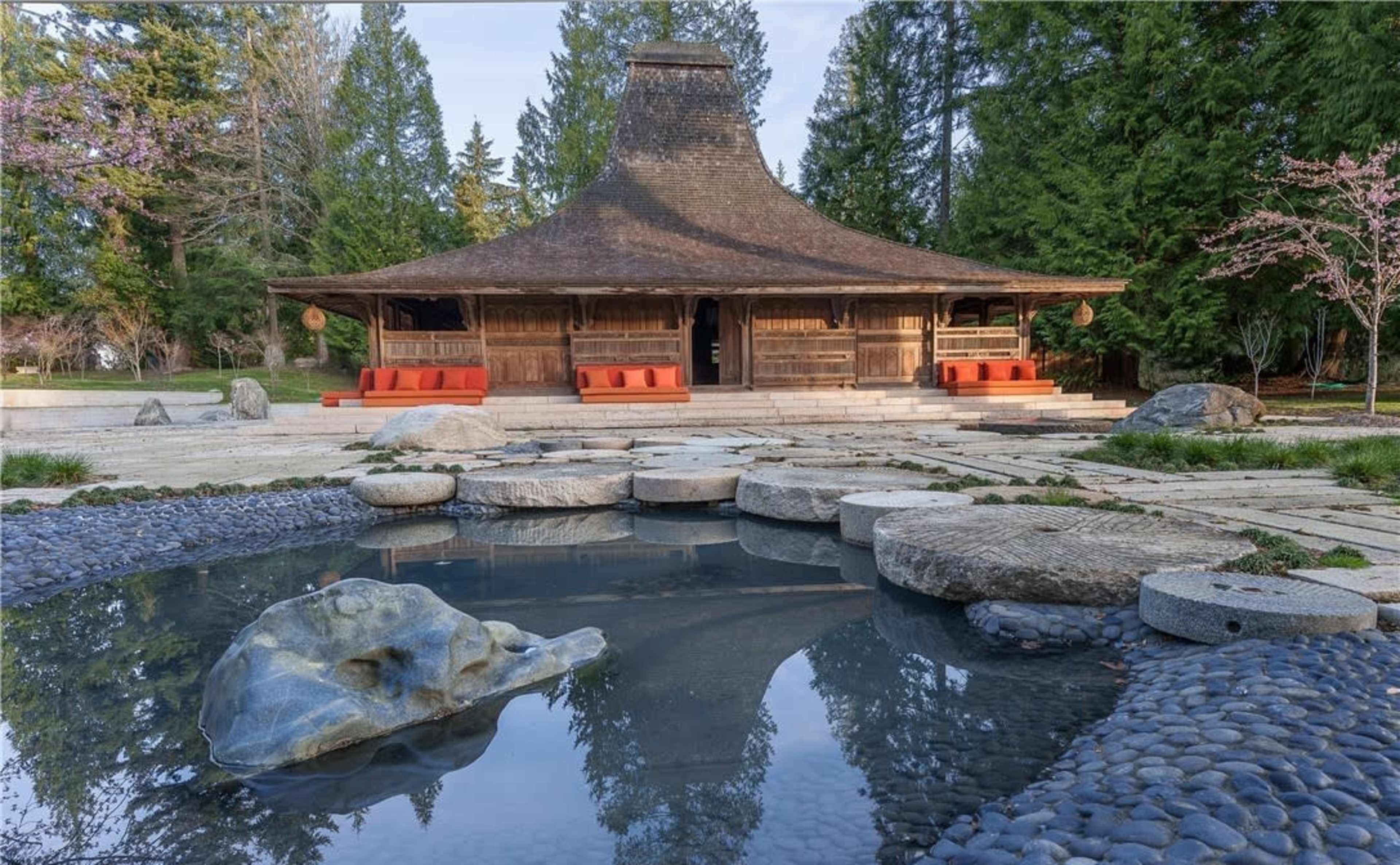 A wooden building with a unique roof design is surrounded by a stone pathway, a pond reflecting the structure, and landscaped greenery.