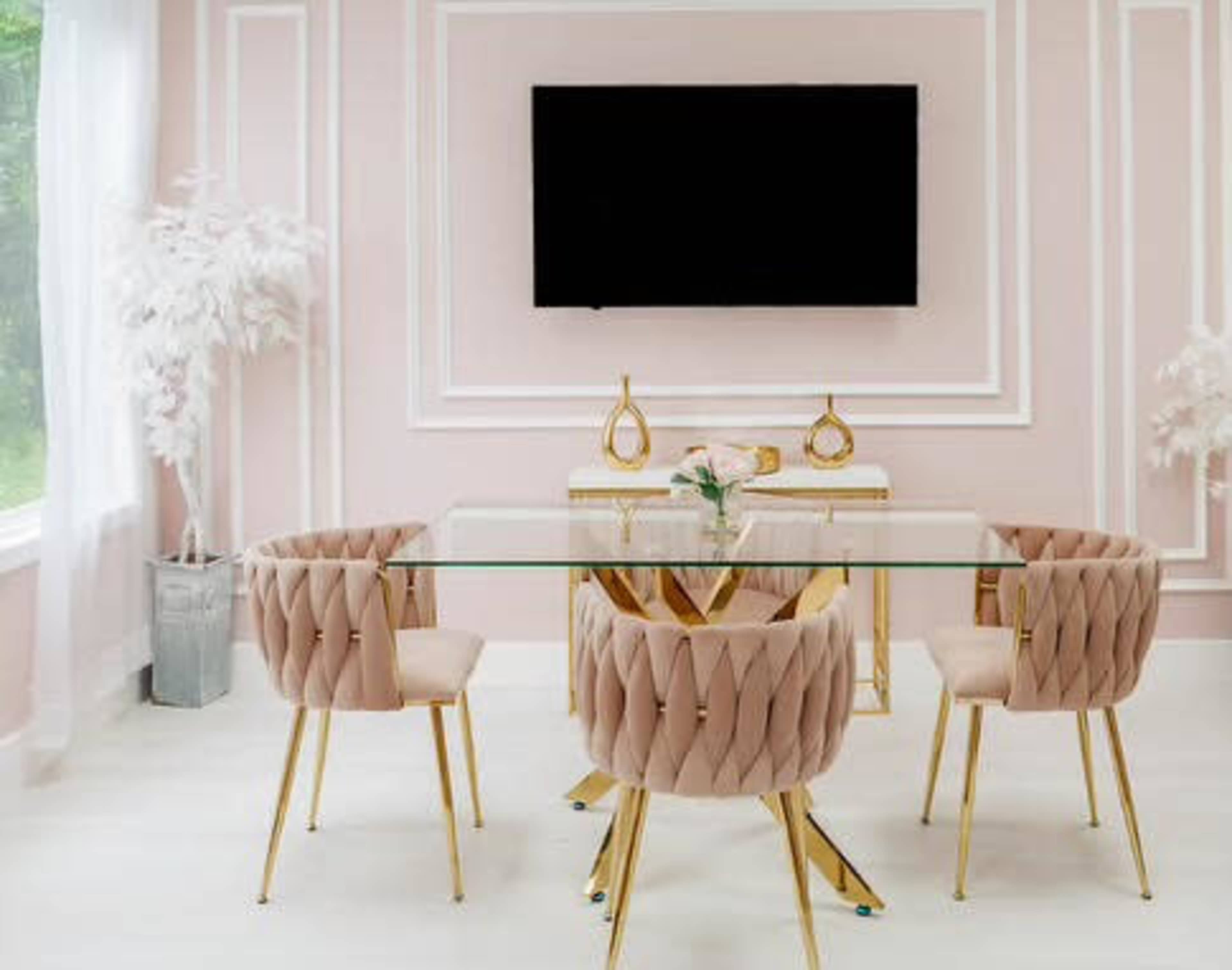 A modern dining room features a glass table surrounded by four blush pink chairs with gold legs, against a pale pink wall with a mounted television above.