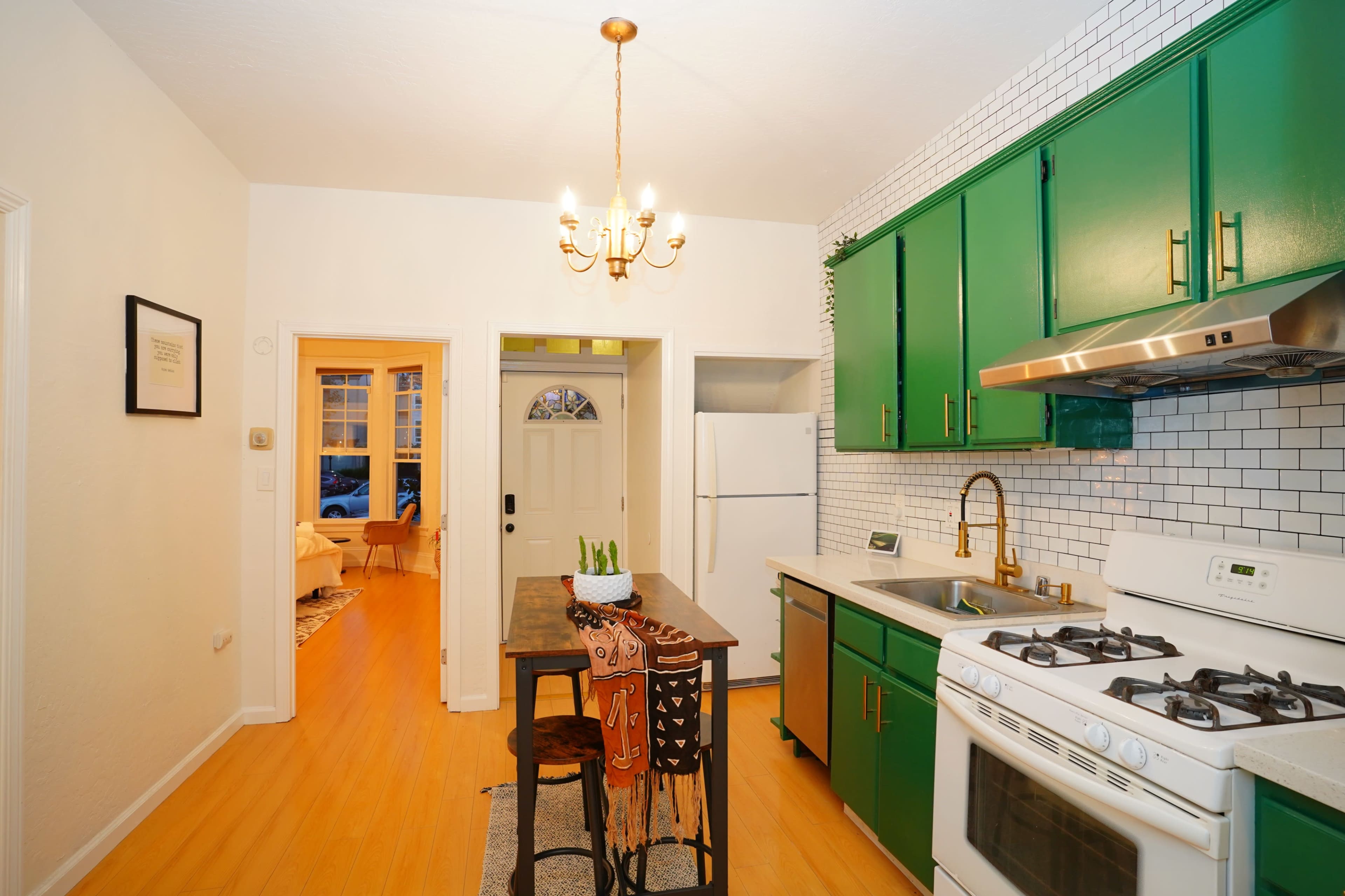 A bright kitchen features green cabinets, white tiled walls, and a small dining table with two chairs, with an entrance leading to another room.