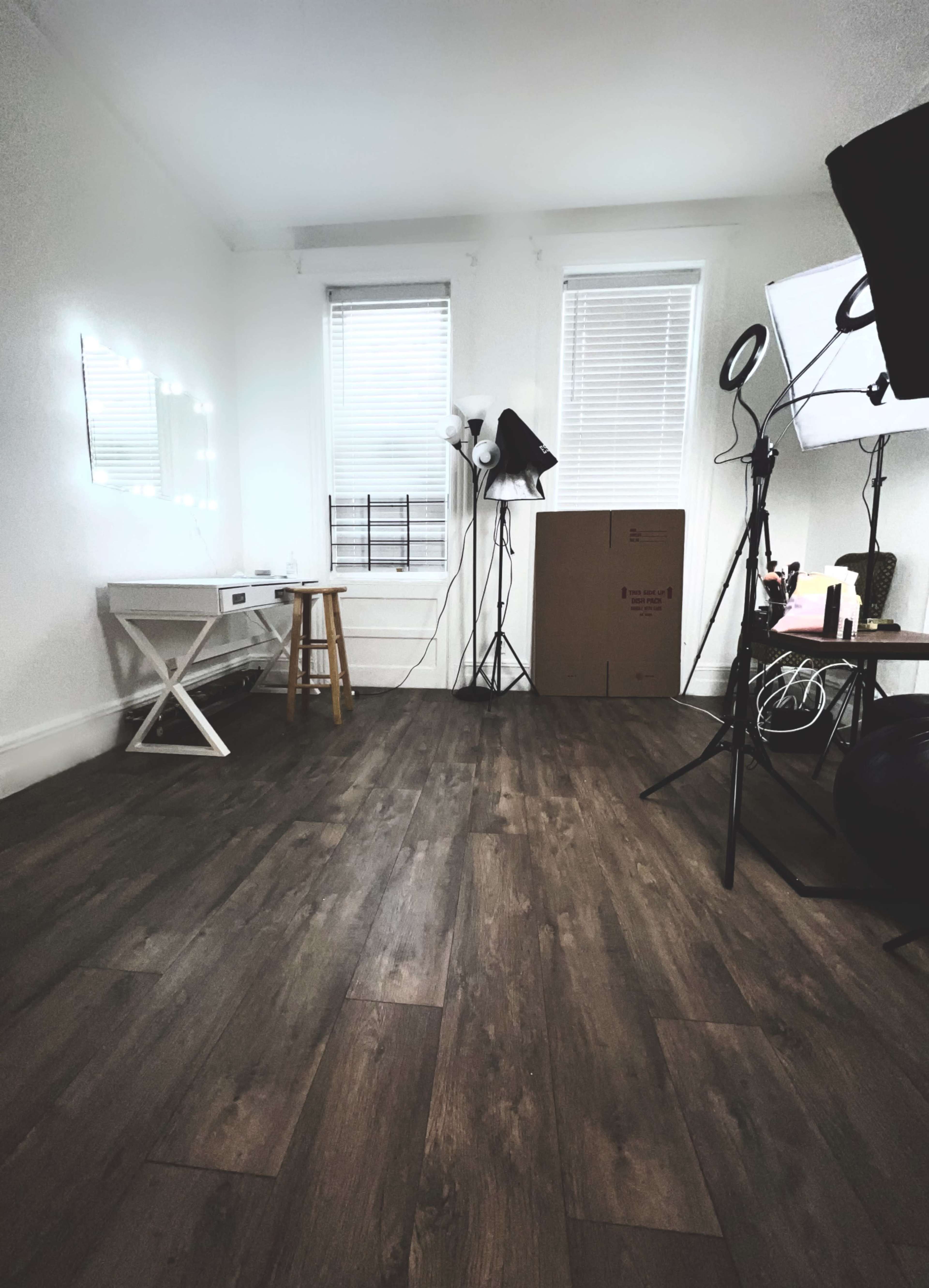 A well-lit room features a white table with a stool, a cardboard box, a chair, and studio lighting equipment against the walls.