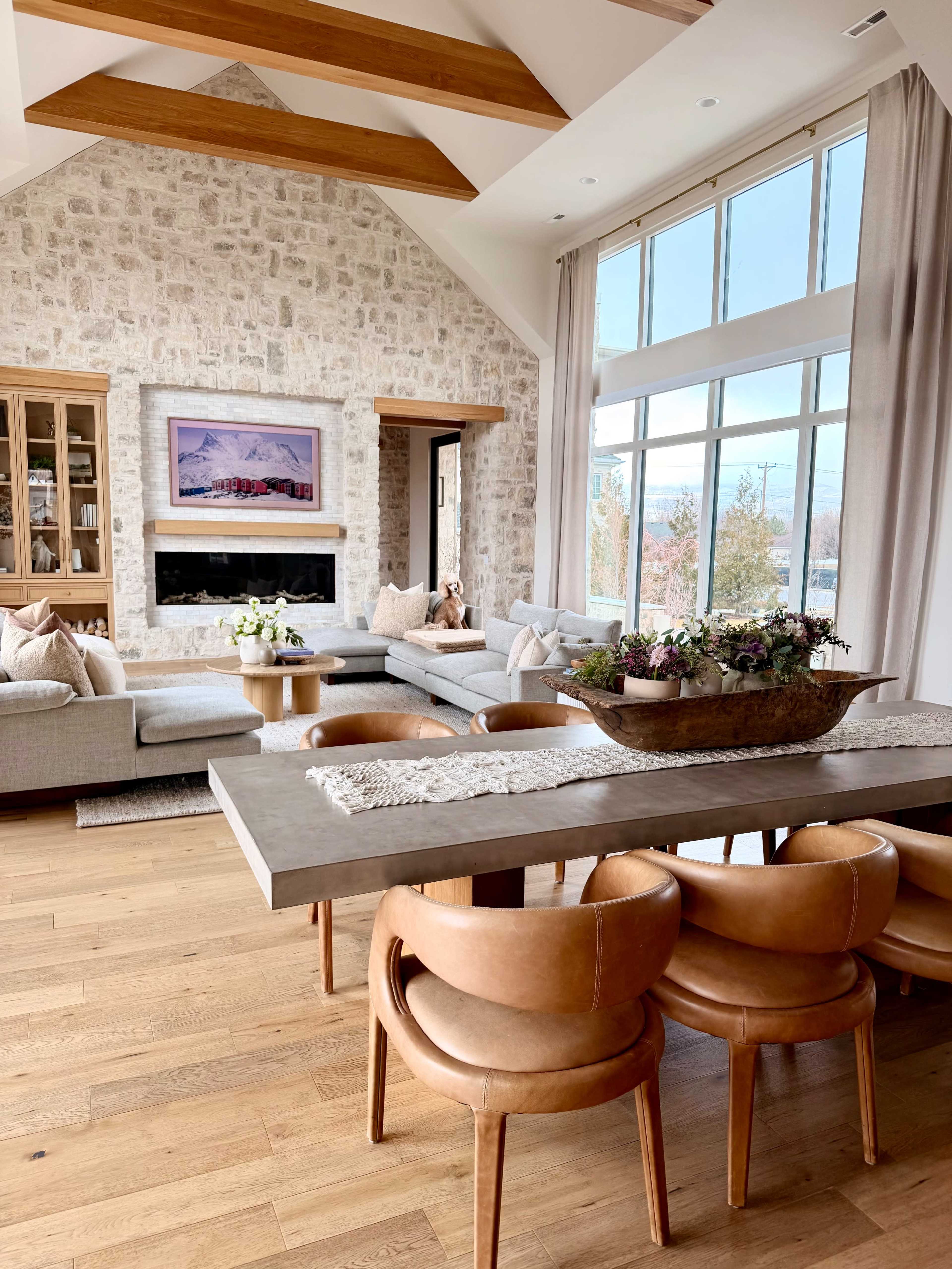 The spacious living room features a stone wall with a fireplace, large windows letting in natural light, and a dining area with a wooden table surrounded by curved leather chairs.