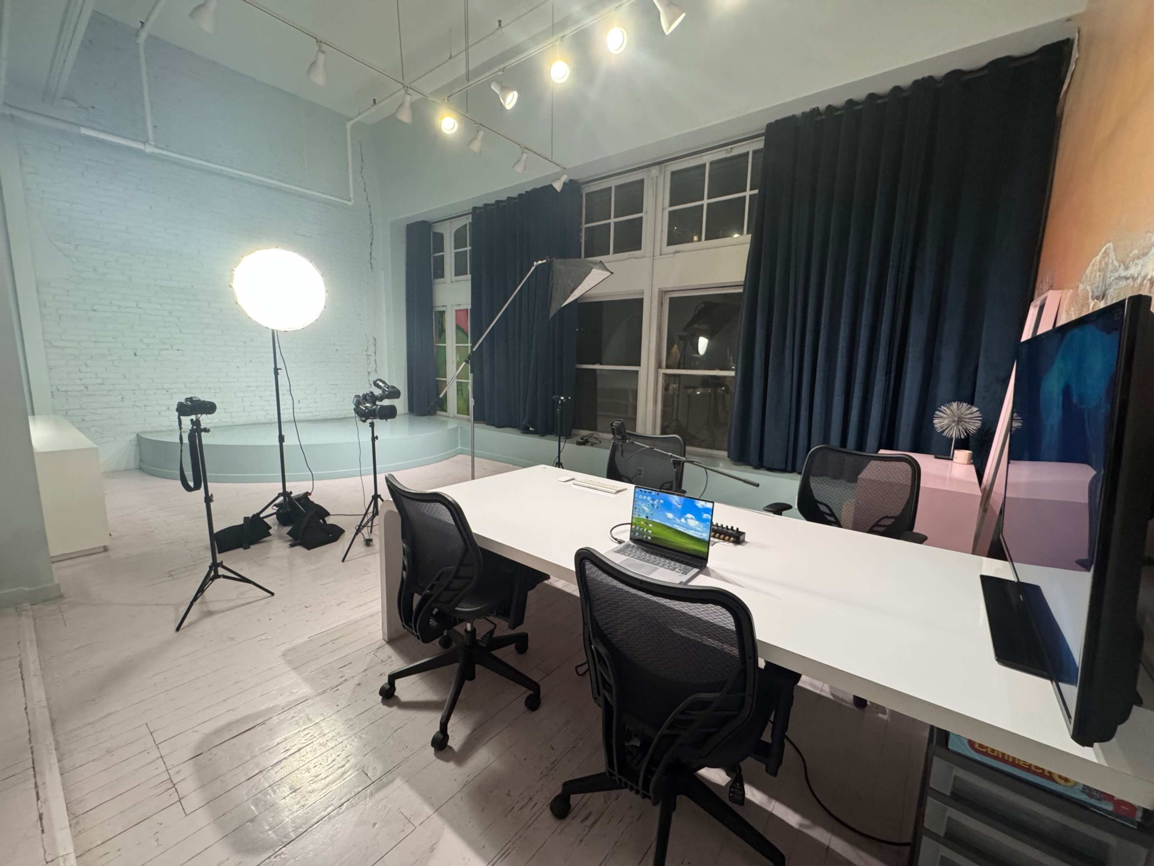 The image shows a modern workspace with a long white desk, several cameras on tripods, a large lamp, and dark curtains covering the windows.