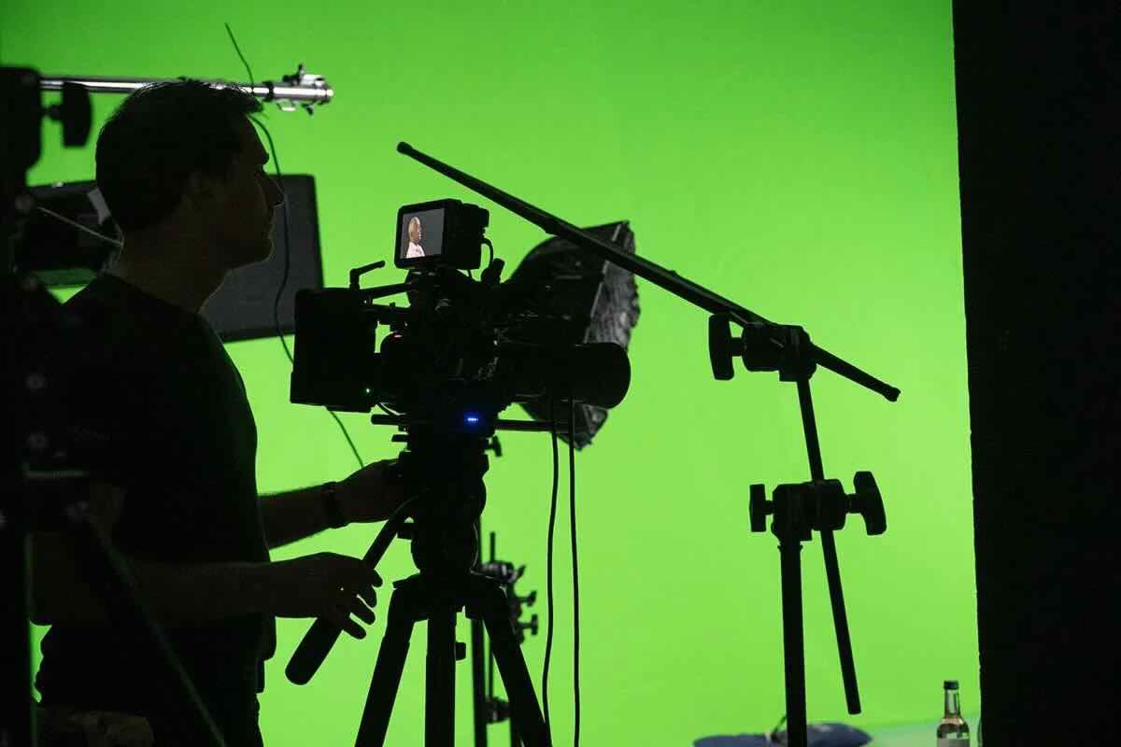 A person operates a camera on a tripod in front of a bright green backdrop.