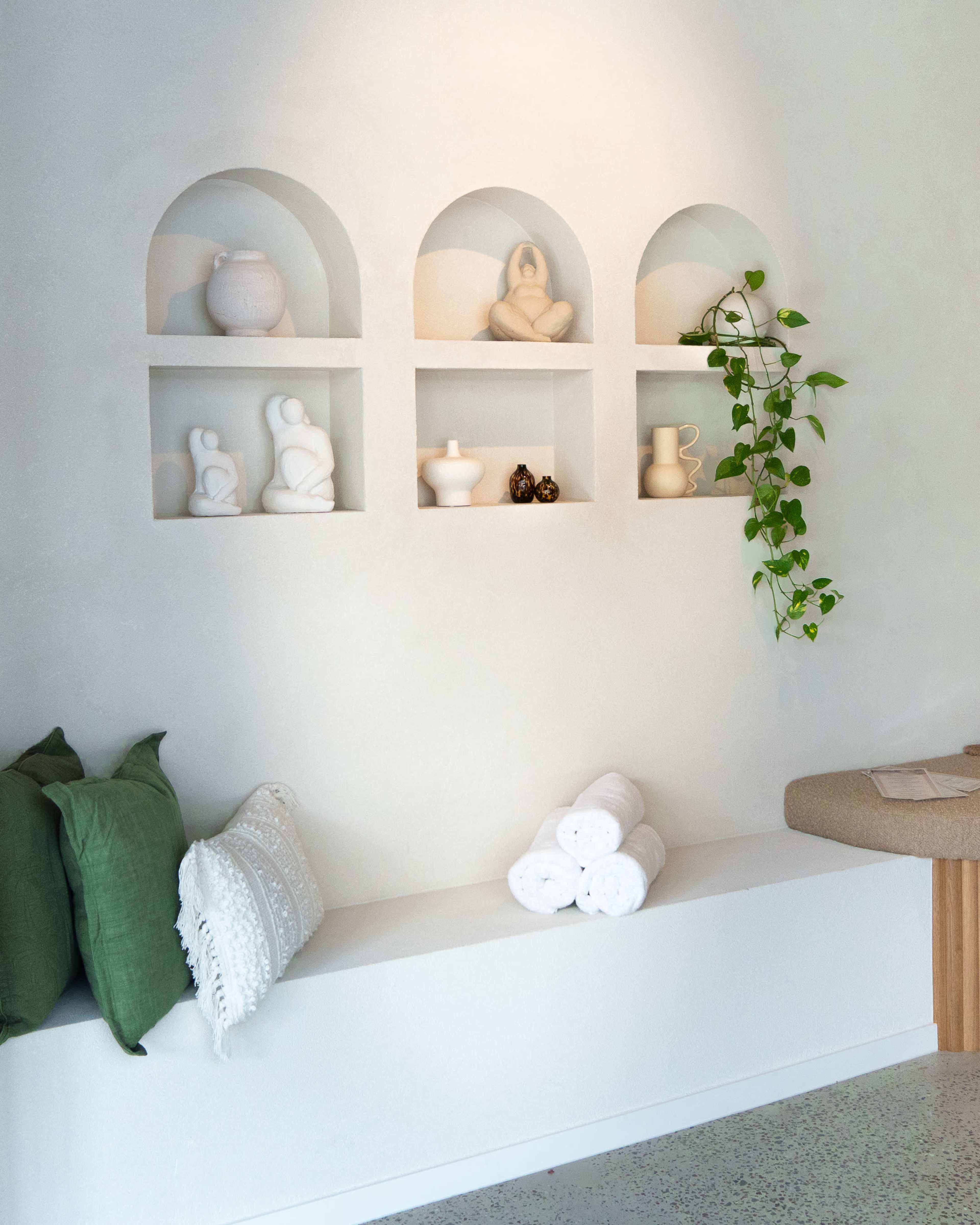 The image features a minimalist interior with three arch-shaped shelves displaying ceramic sculptures and a potted plant, accompanied by decorative pillows and rolled towels on a bench.