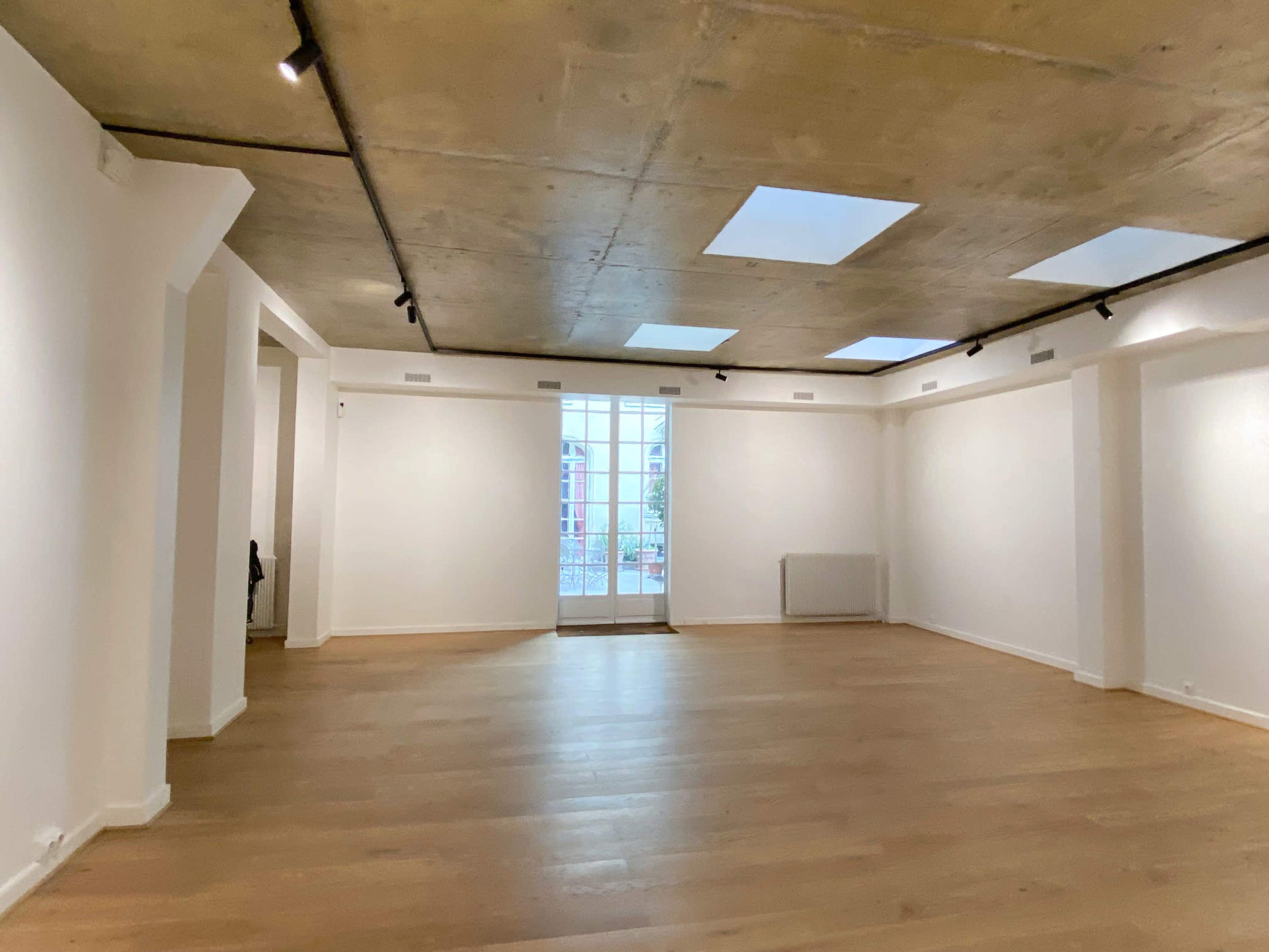 The image shows an empty art gallery with a concrete ceiling, wall-mounted lights, and large windows at one end.