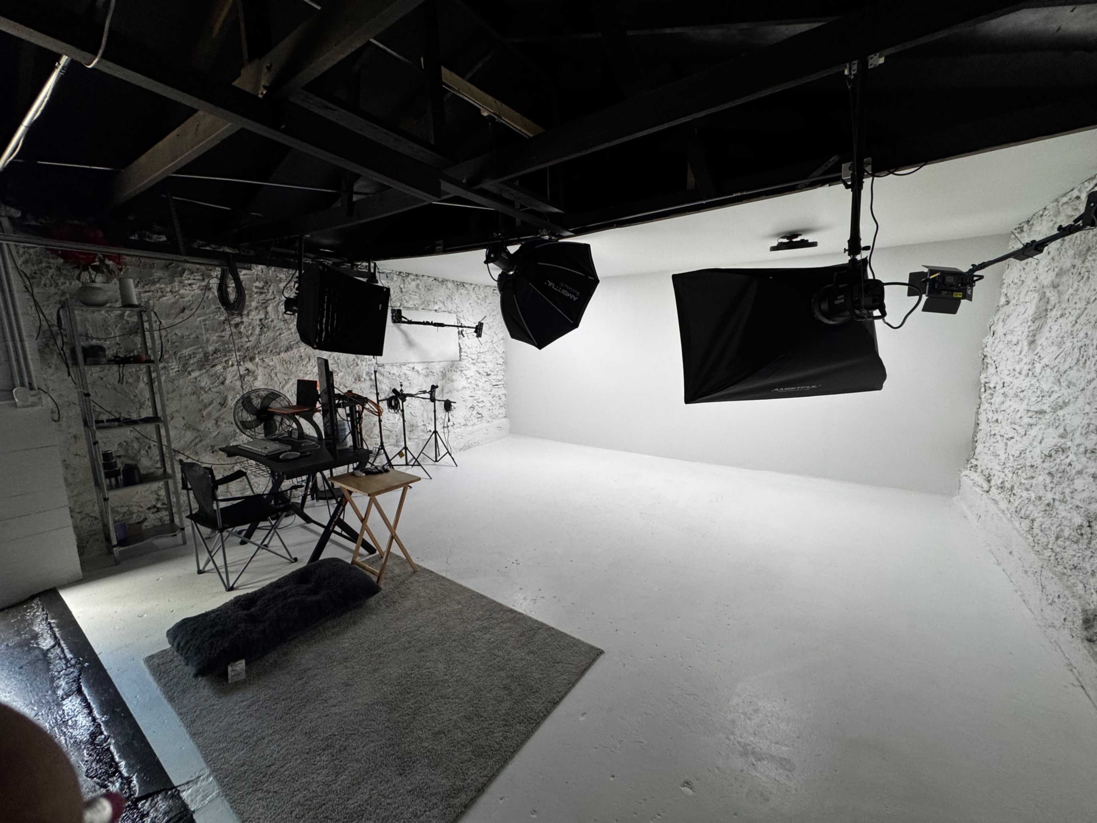 The image shows a bright, empty photography studio with various lighting equipment, chairs, and a rug on the floor.