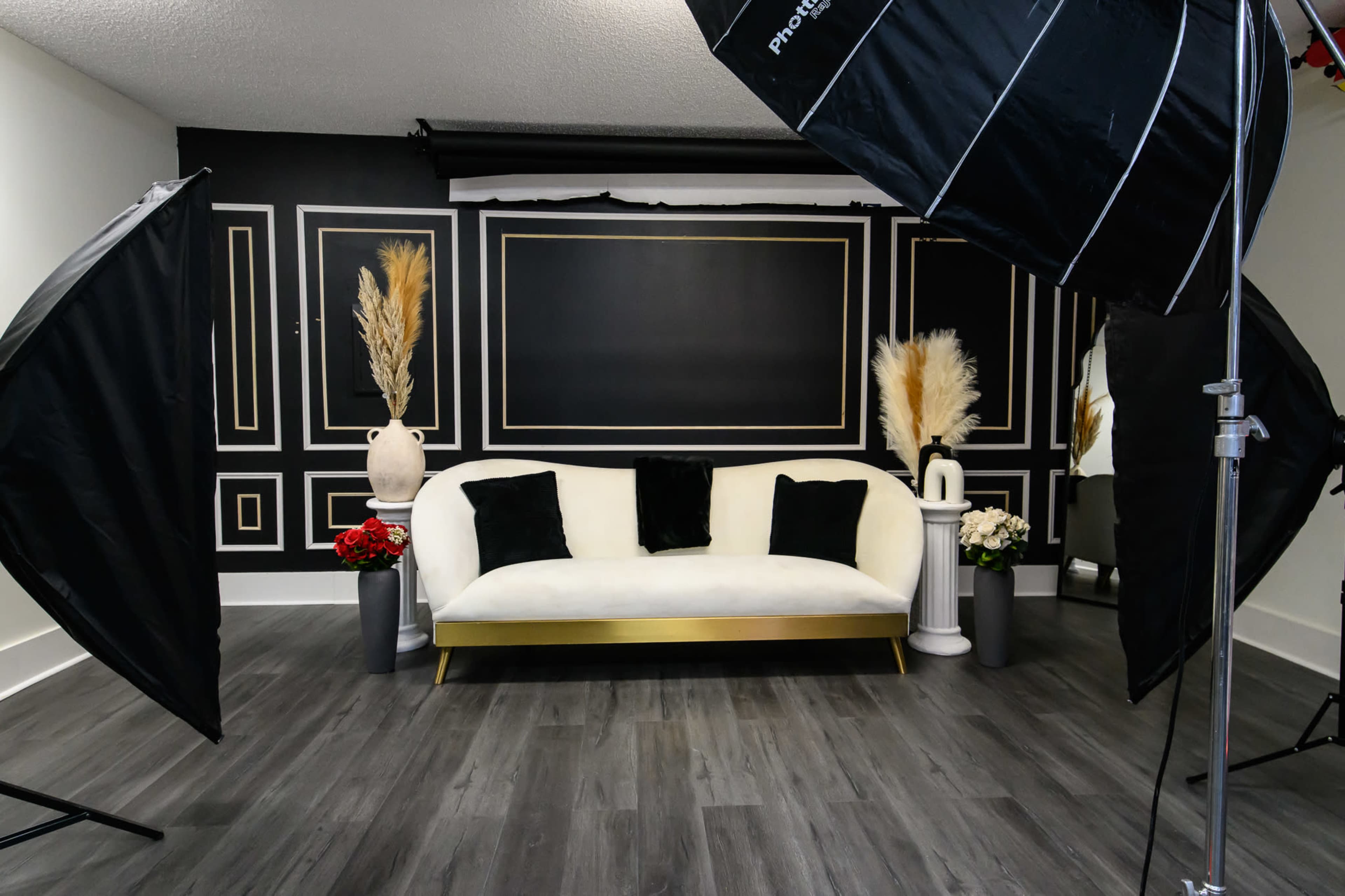 A well-lit photography studio featuring a white sofa with black pillows, flanked by large softbox lights and decorative vases with plants.