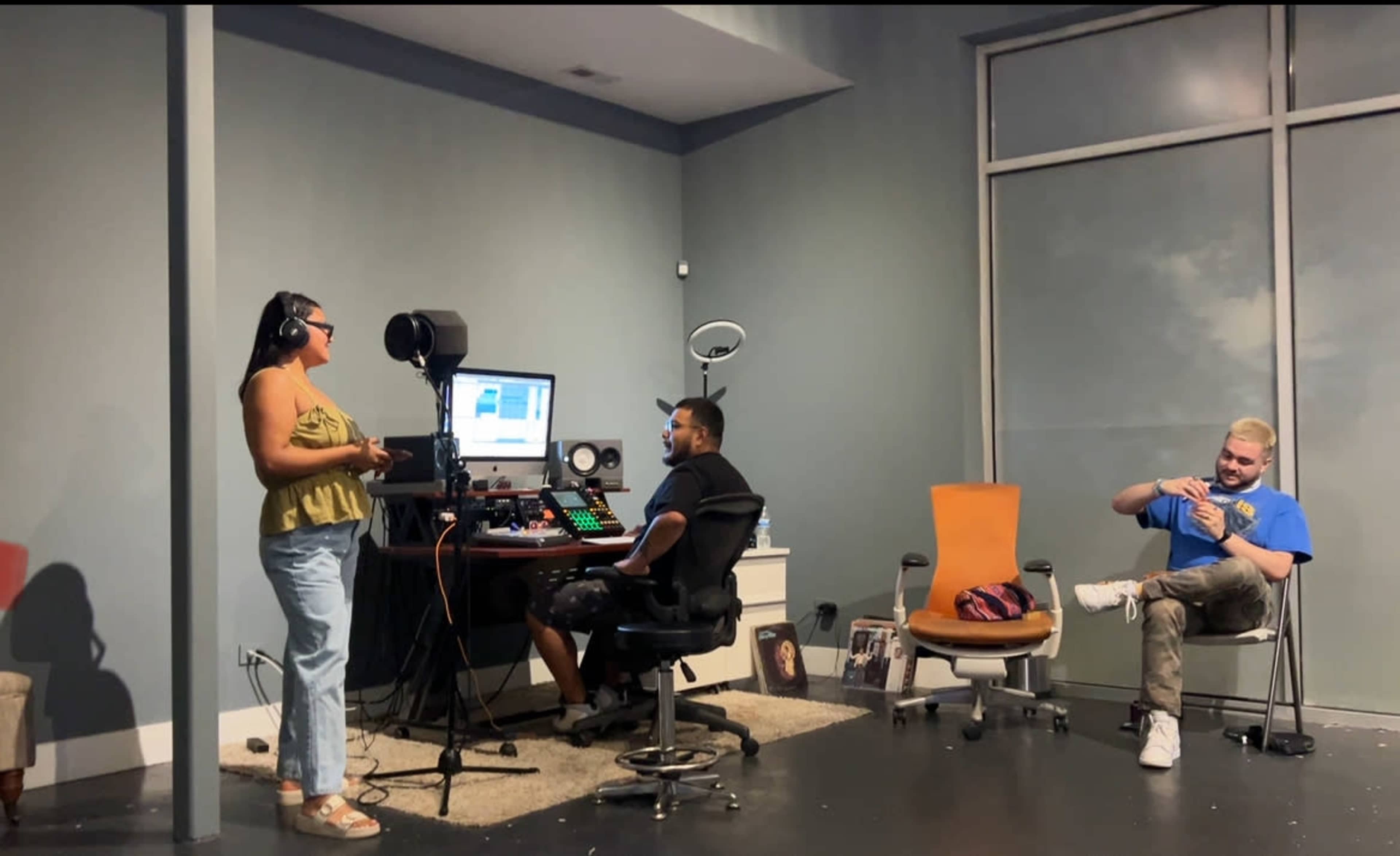A woman stands in front of a microphone while a man sits at a computer with audio equipment, and another man relaxes in an orange chair nearby.