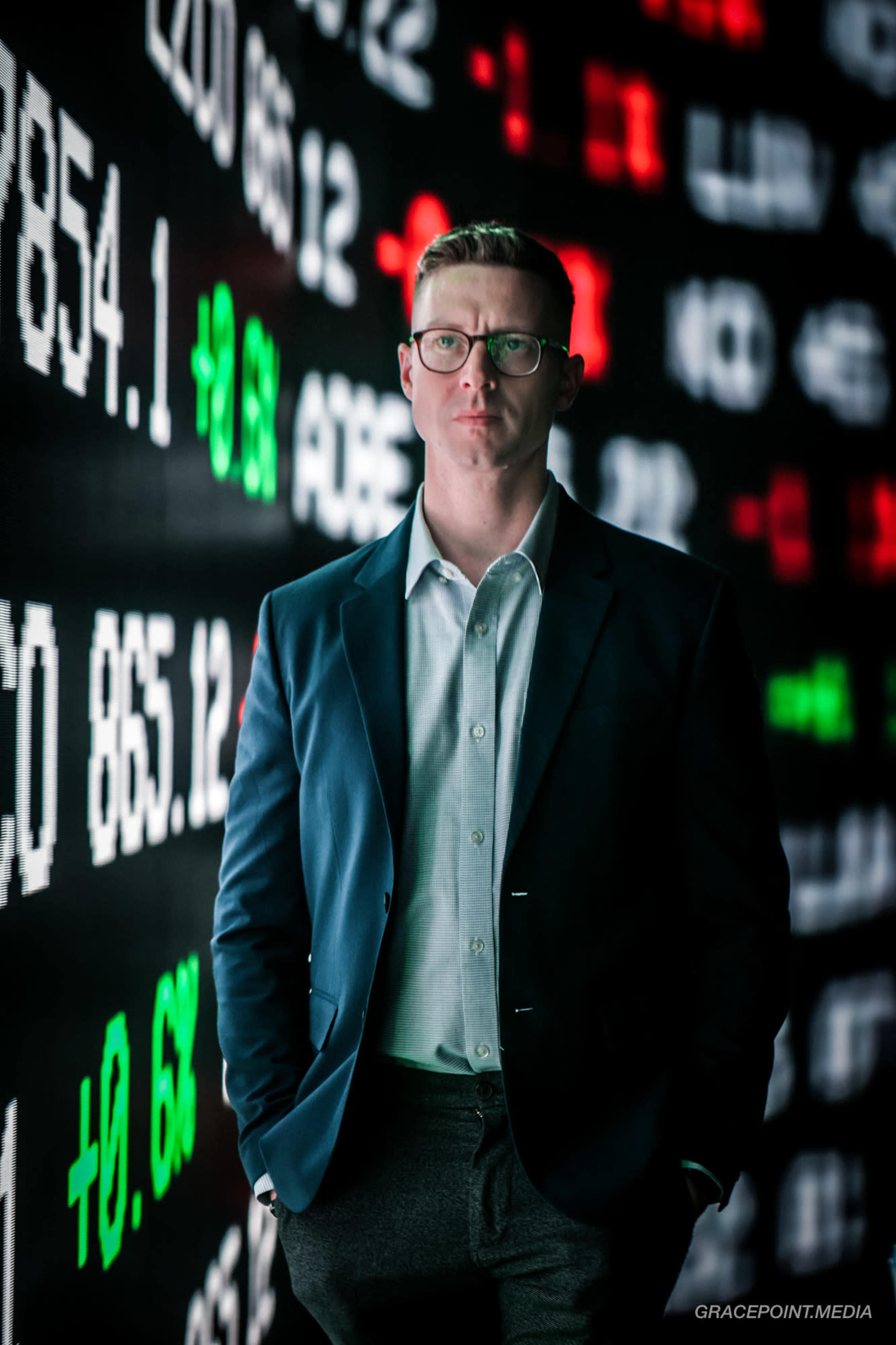 A man in glasses stands in front of a digital display of stock market data.