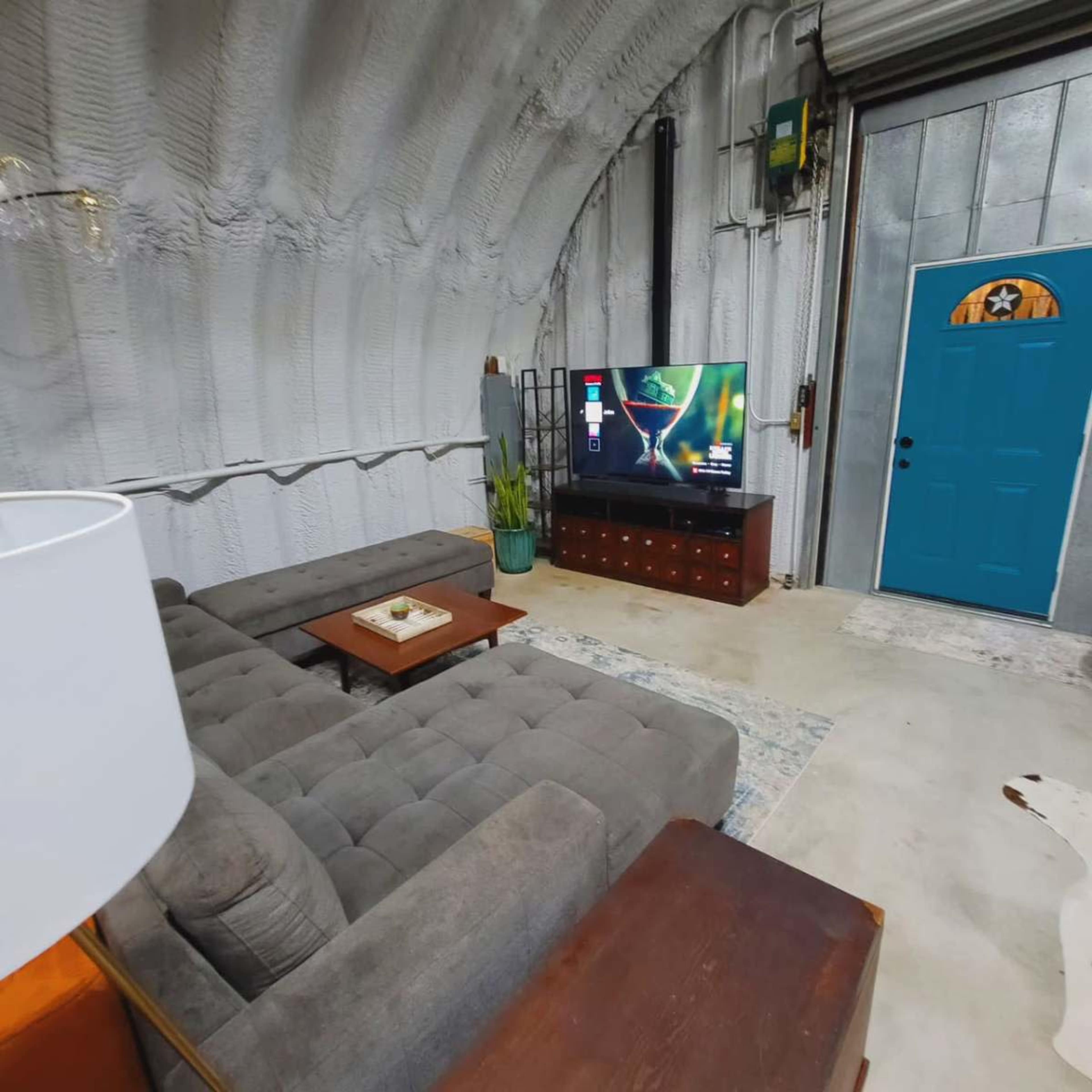 A cozy living room with a gray sectional sofa, a wooden coffee table, a TV mounted on the wall, and a blue door leading outside.