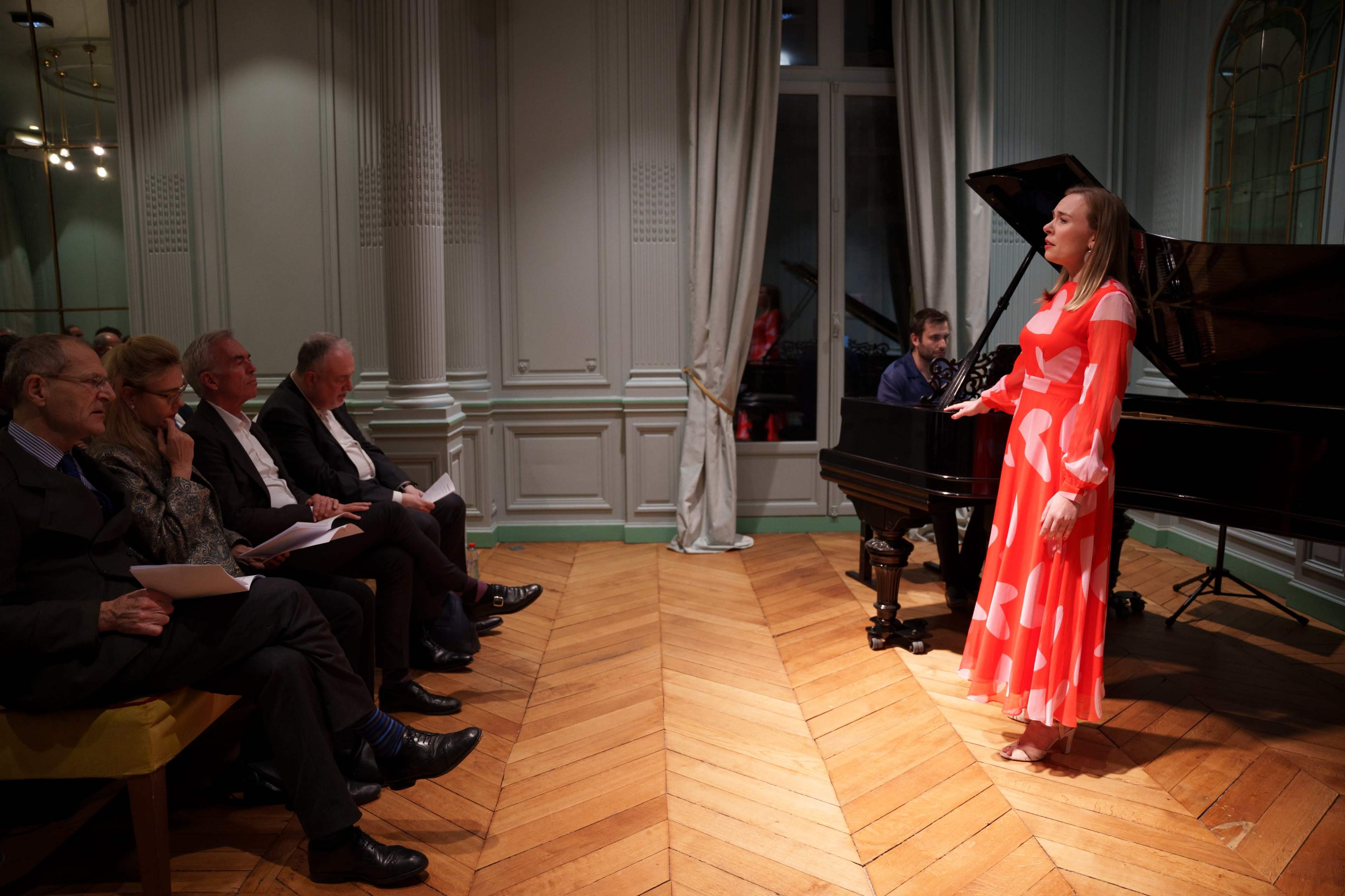 A woman in a flowy orange and white dress sings on stage while a pianist plays, and an audience watches attentively from their seats.