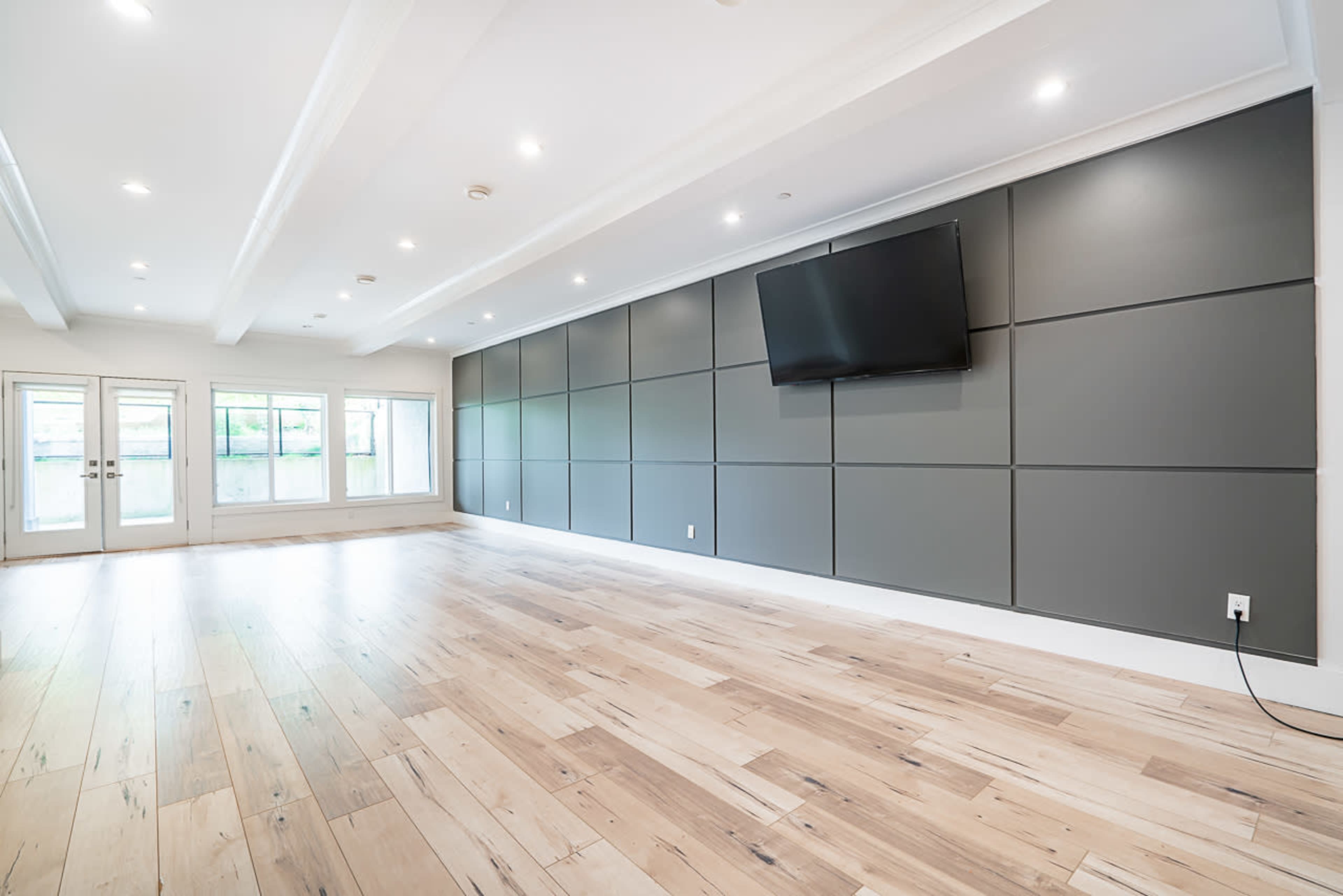 A spacious room with wooden flooring, a gray accent wall, and a mounted television, illuminated by recessed lighting.