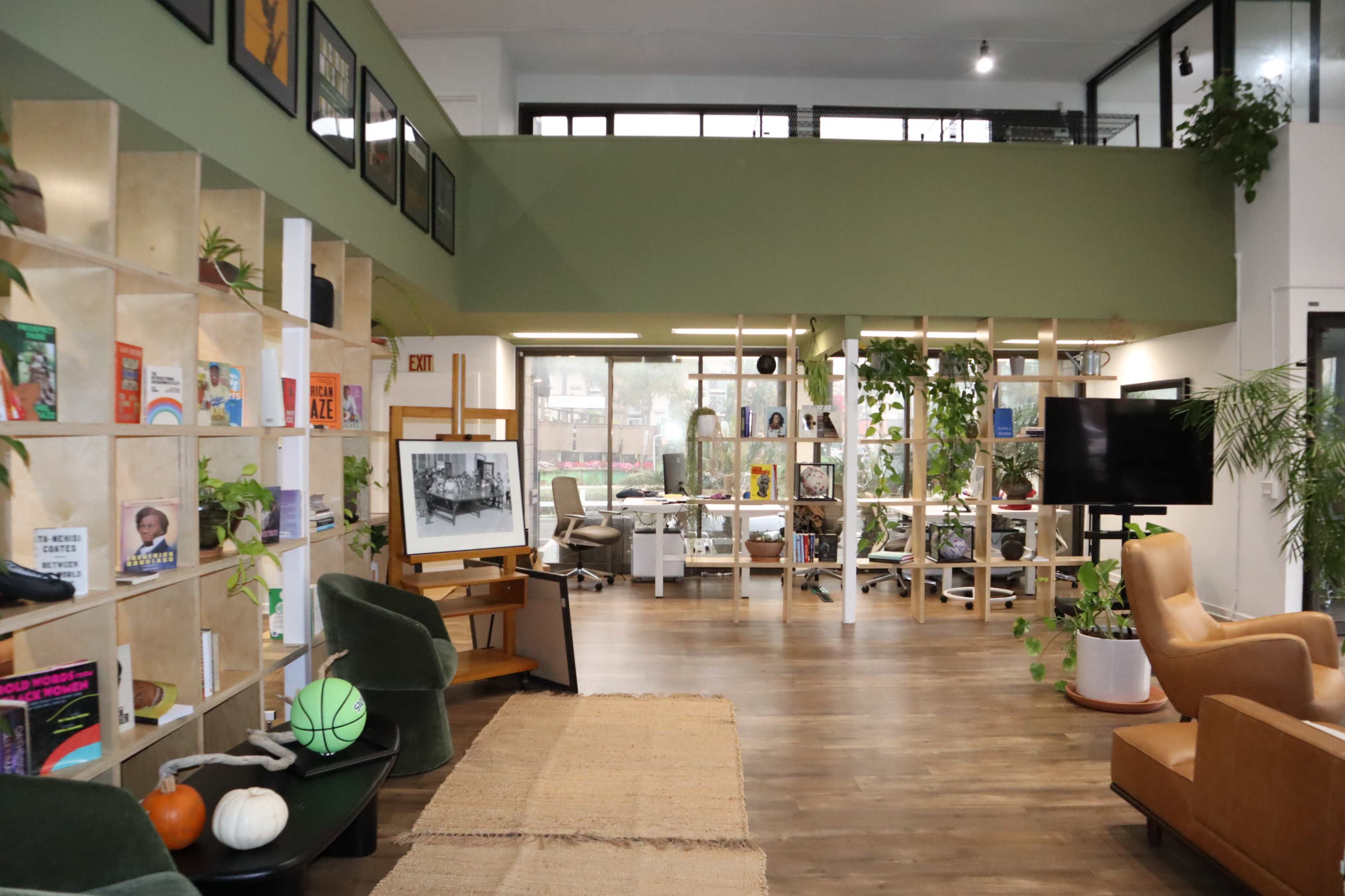 The image shows a modern, open-plan workspace with green walls, plants, and a mix of seating areas and bookshelves.