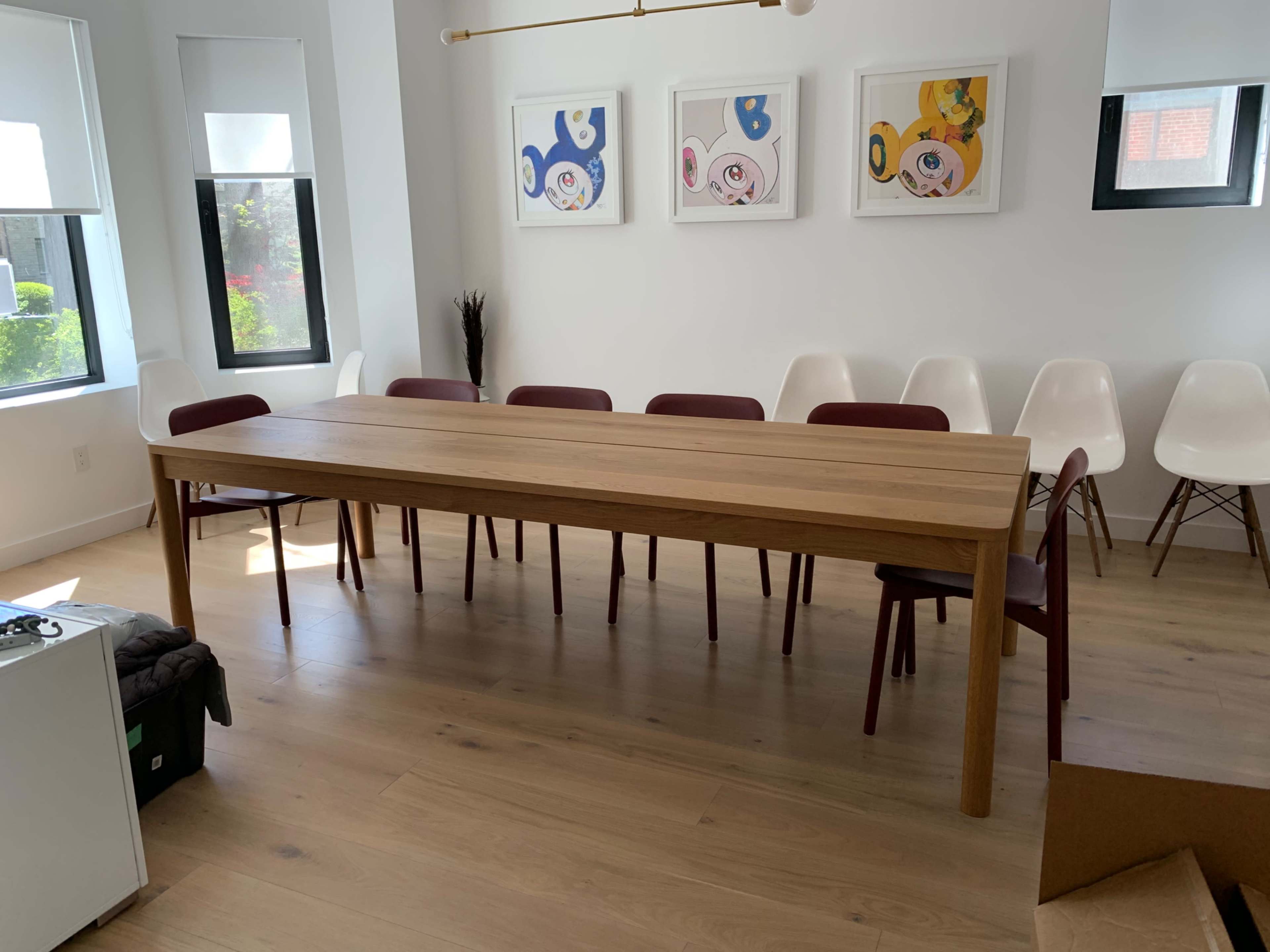 A long wooden dining table is placed in a bright room with several empty chairs arranged around it and framed artworks on the walls.