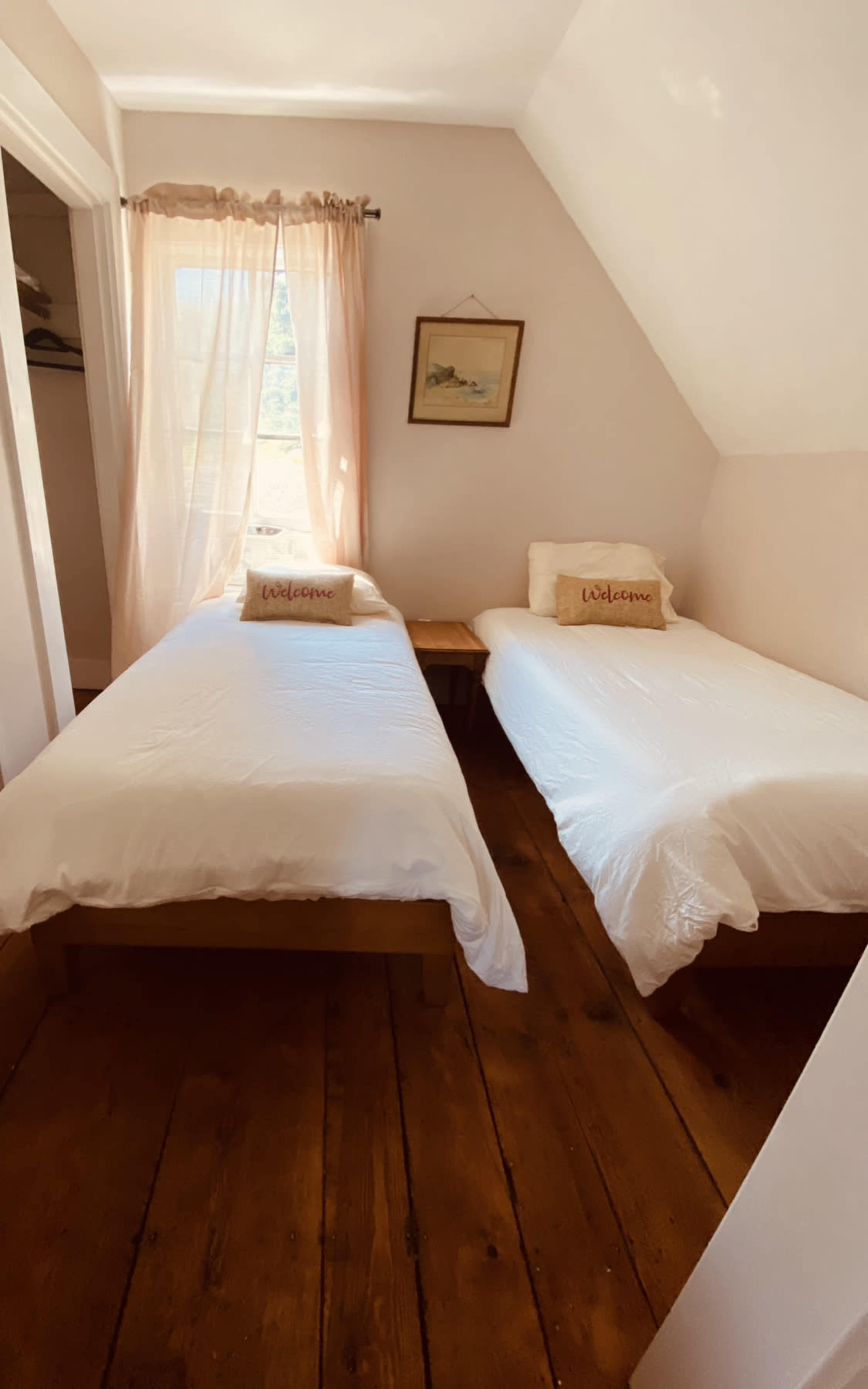A small room with two single beds, each covered with white bedding, and a window with sheer curtains allowing natural light to enter.