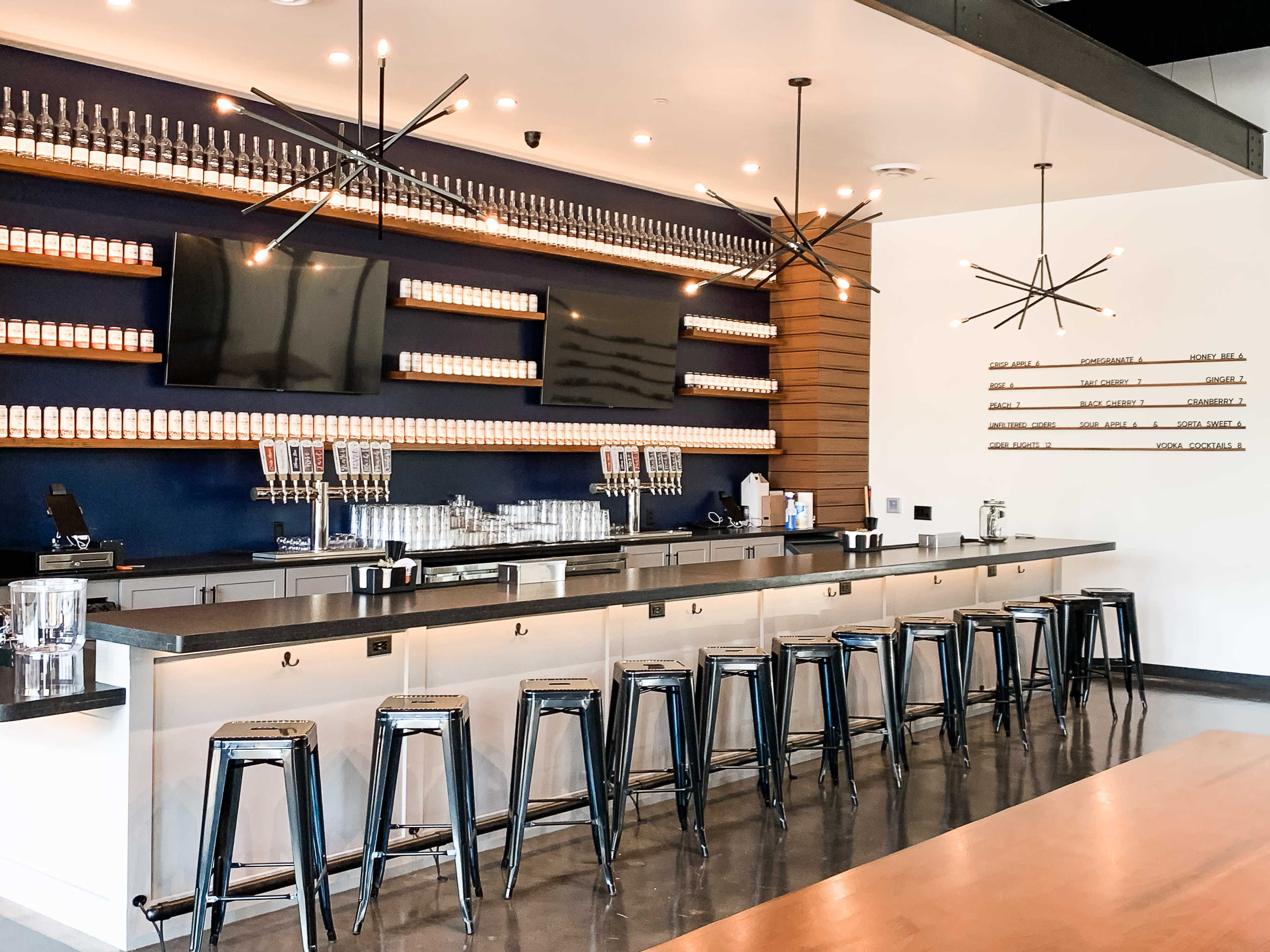 The image shows a modern bar with a sleek counter, metal stools, and a wall of beer taps and bottles in a well-lit setting.