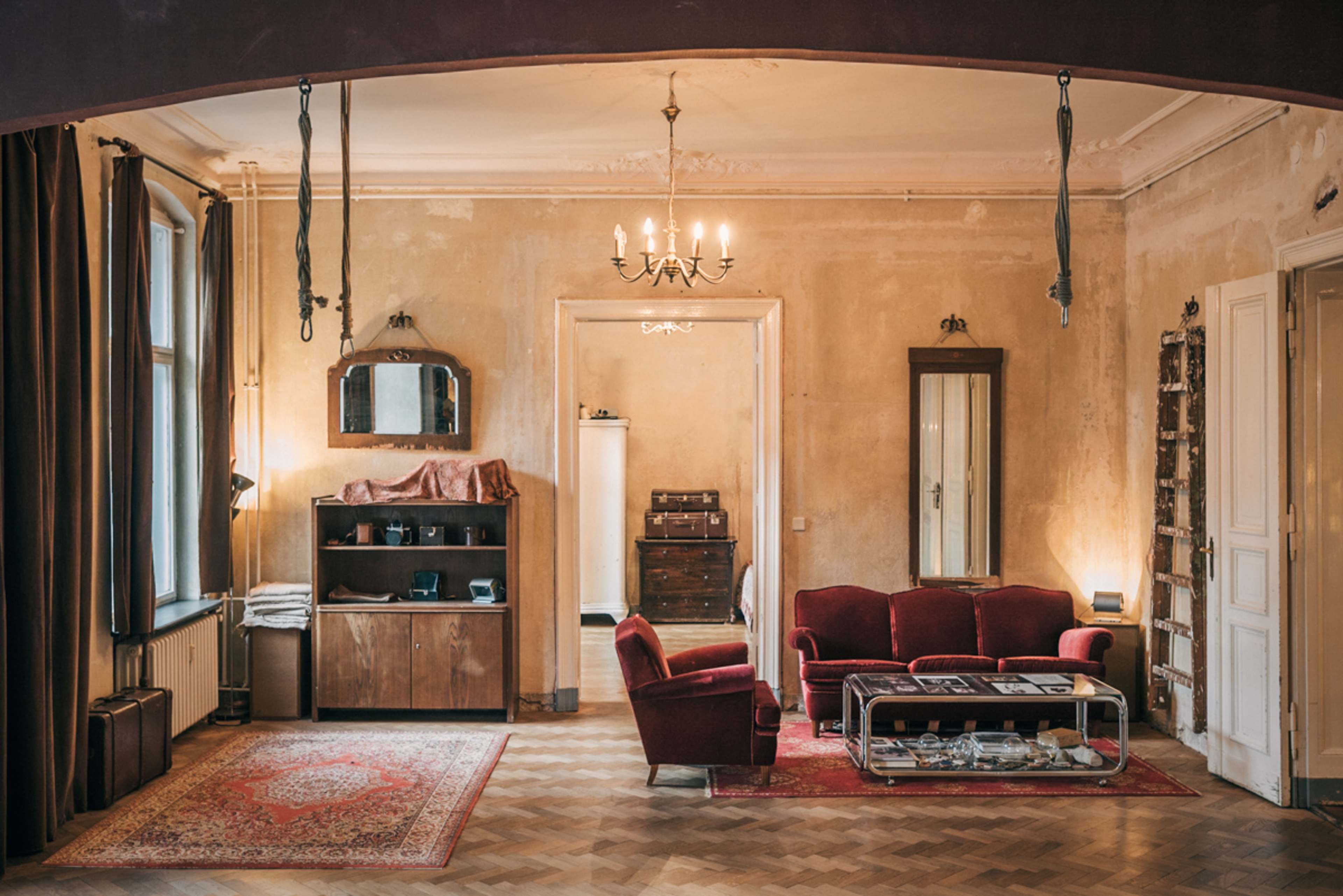 The image shows a vintage-style living room with a red sofa, a small coffee table, and wooden furniture against peeling walls and wooden flooring.