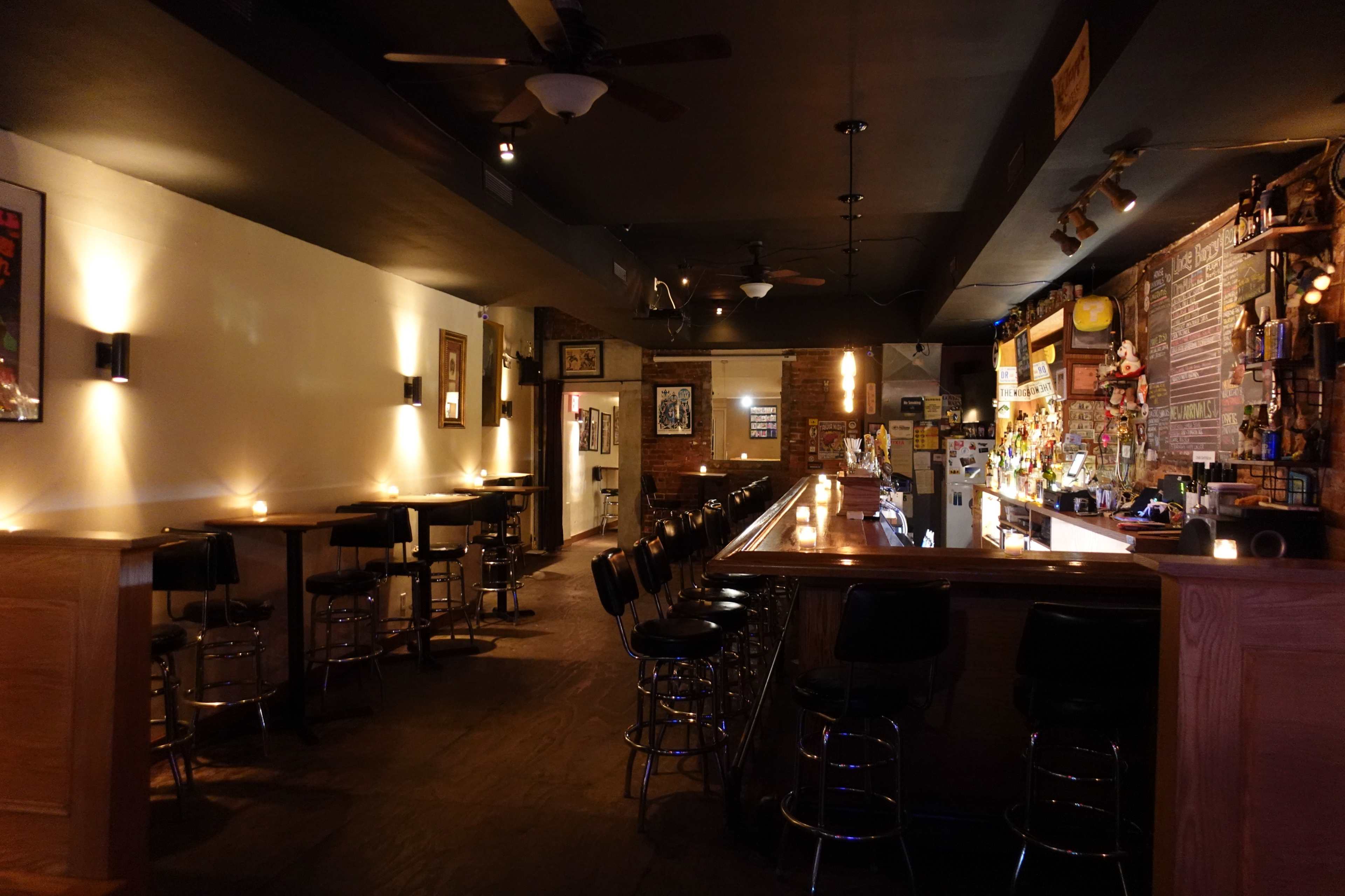 A dimly lit bar with a long counter, several empty stools, and wall decorations, creating a quiet atmosphere.