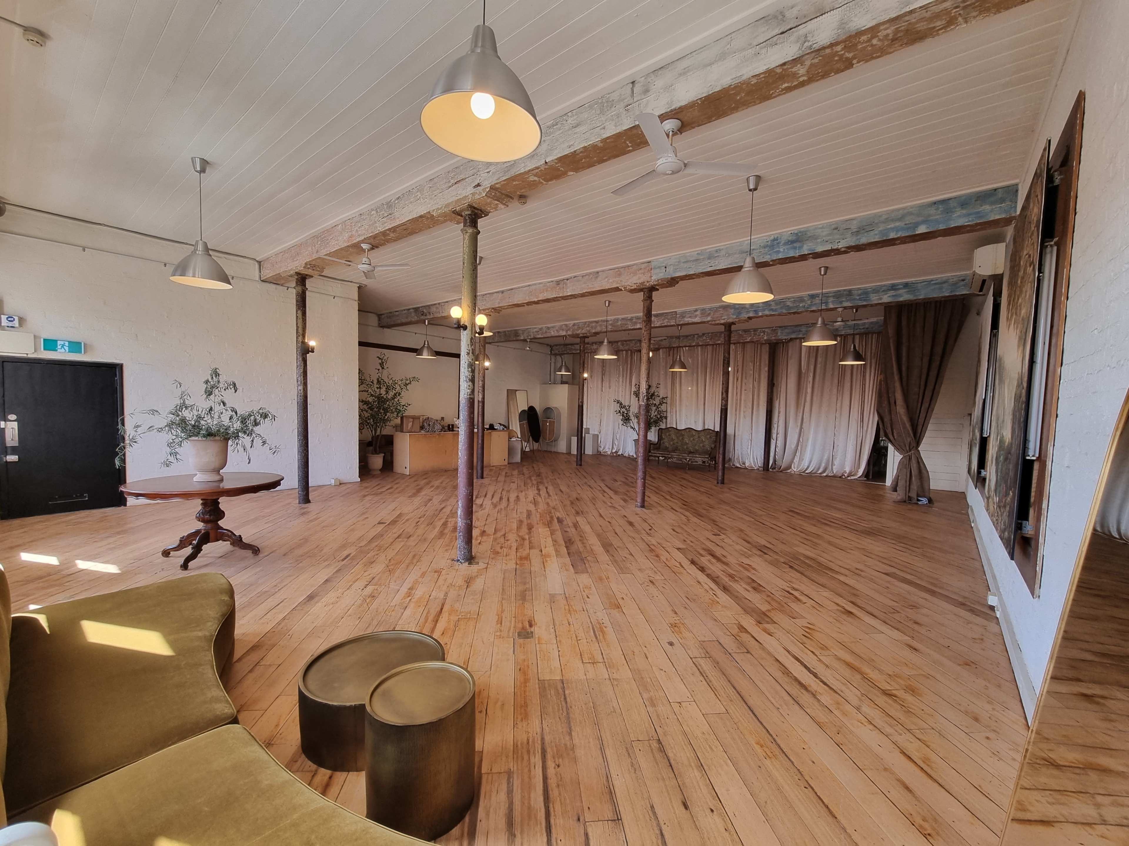 The image shows a spacious room with wooden flooring, exposed beams, and a few plants, featuring a seating area and curtains on one side.