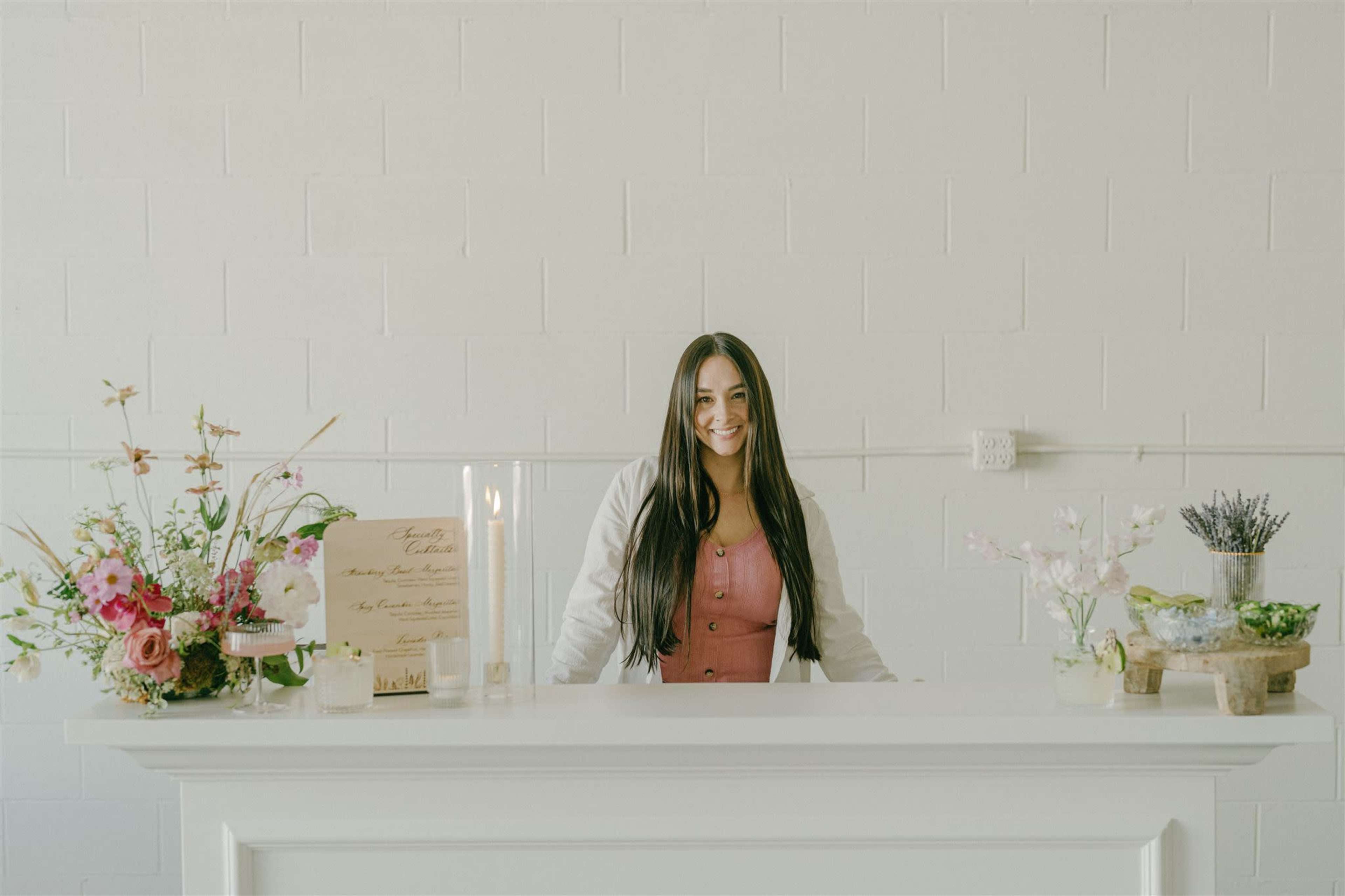 A woman stands behind a white reception desk adorned with floral arrangements and decorative items against a plain wall.