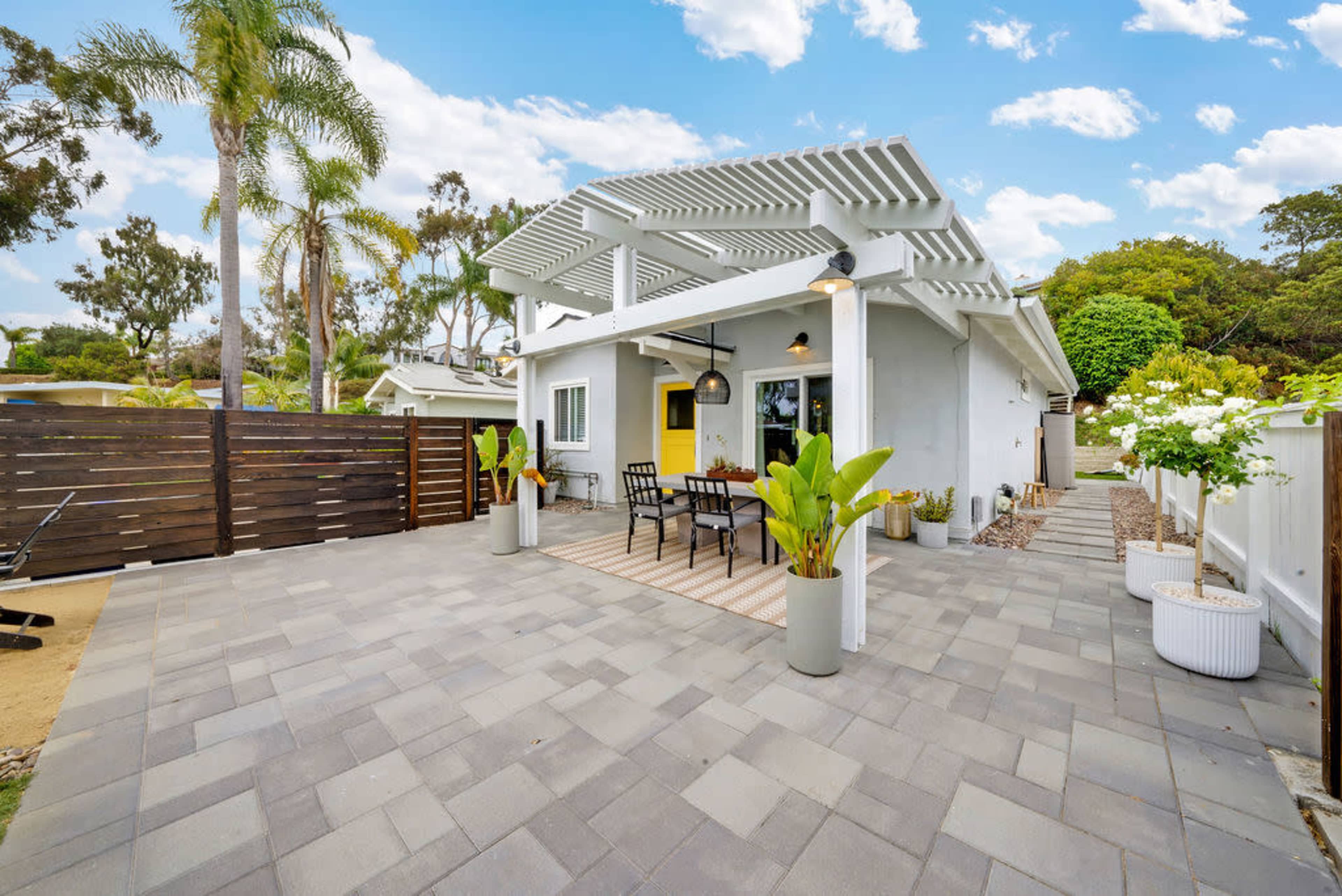 A modern single-story house features a patio with outdoor seating, surrounded by landscaped greenery and a wooden fence.
