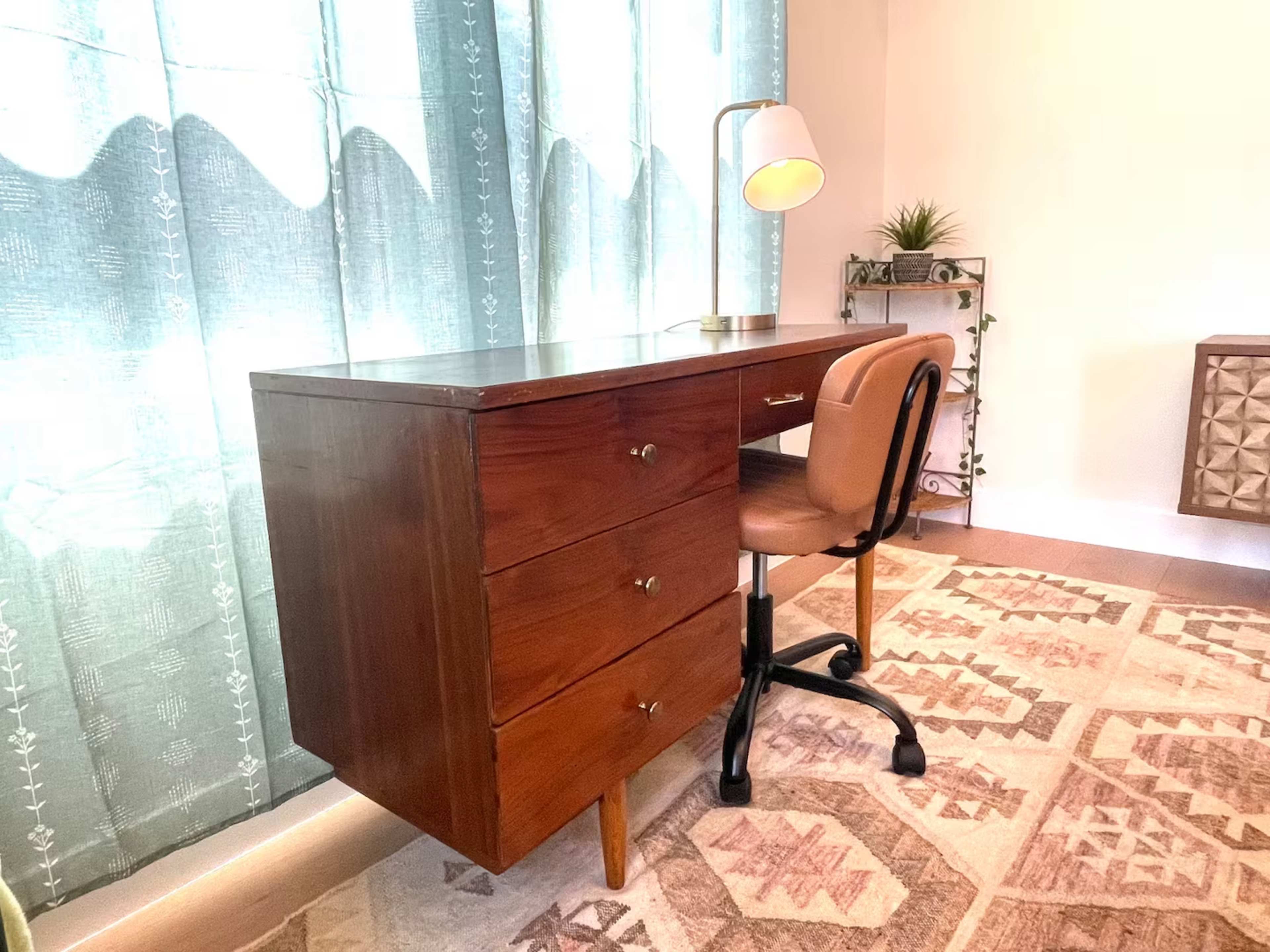 A wooden desk with three drawers and a lamp sits beside a chair on a patterned rug against a wall with light blue curtains.