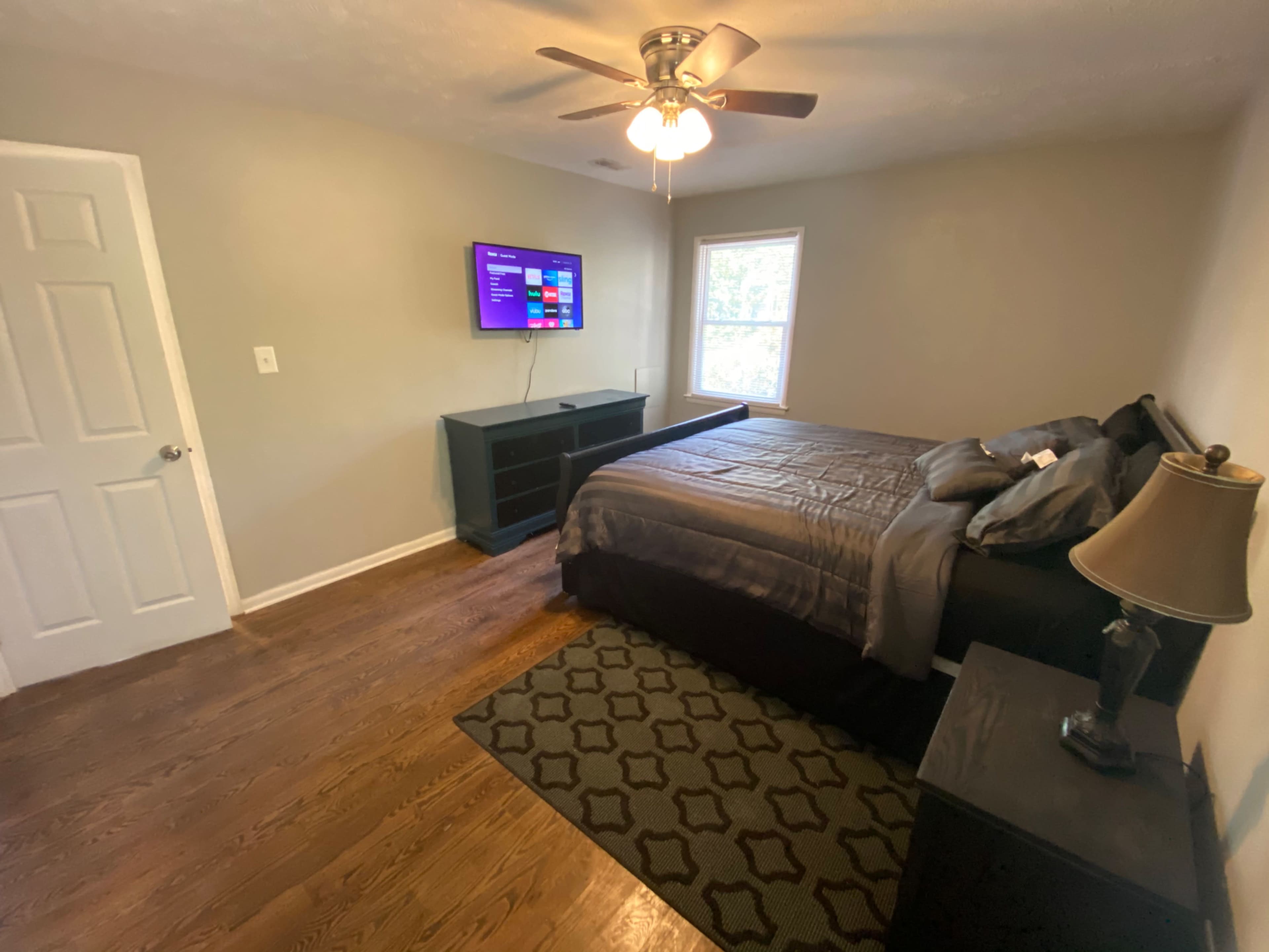 A bedroom with a ceiling fan, a large bed, a TV mounted on the wall, and a wooden floor.