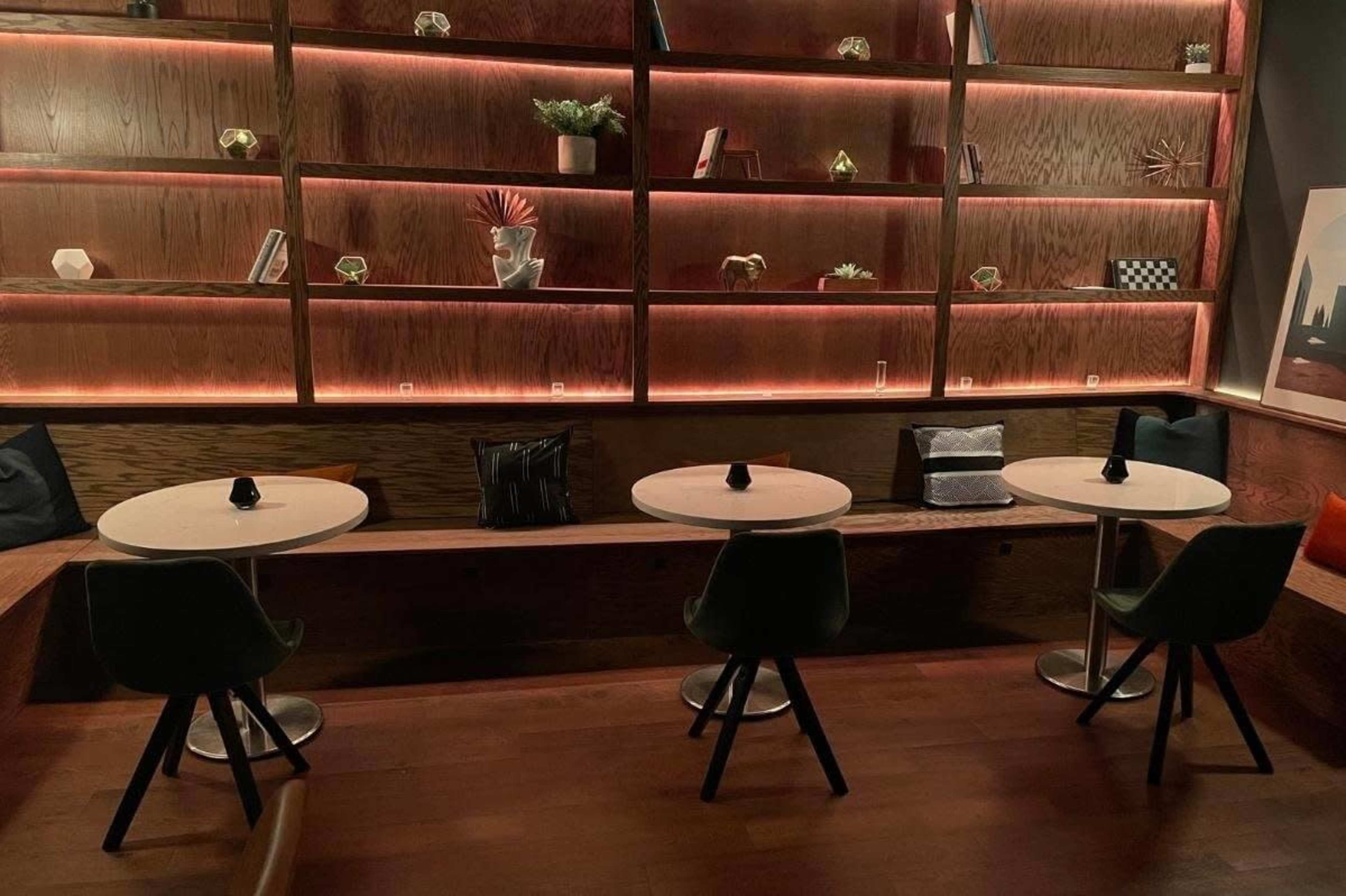The image shows a cozy seating area with three small round tables, each accompanied by a green chair, against a backdrop of illuminated wooden shelves displaying various decorative items.