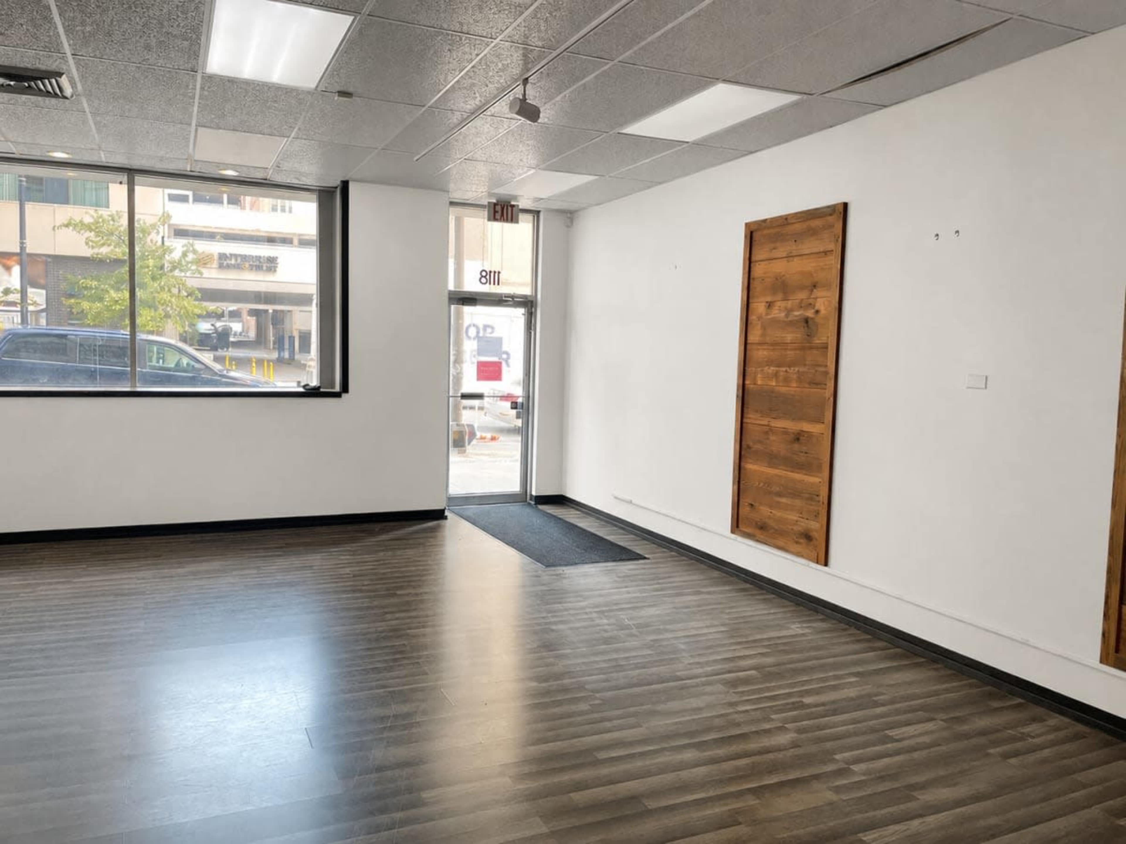 The image shows a spacious, empty commercial space with a view of the street through a large front window and a wooden door partially open.