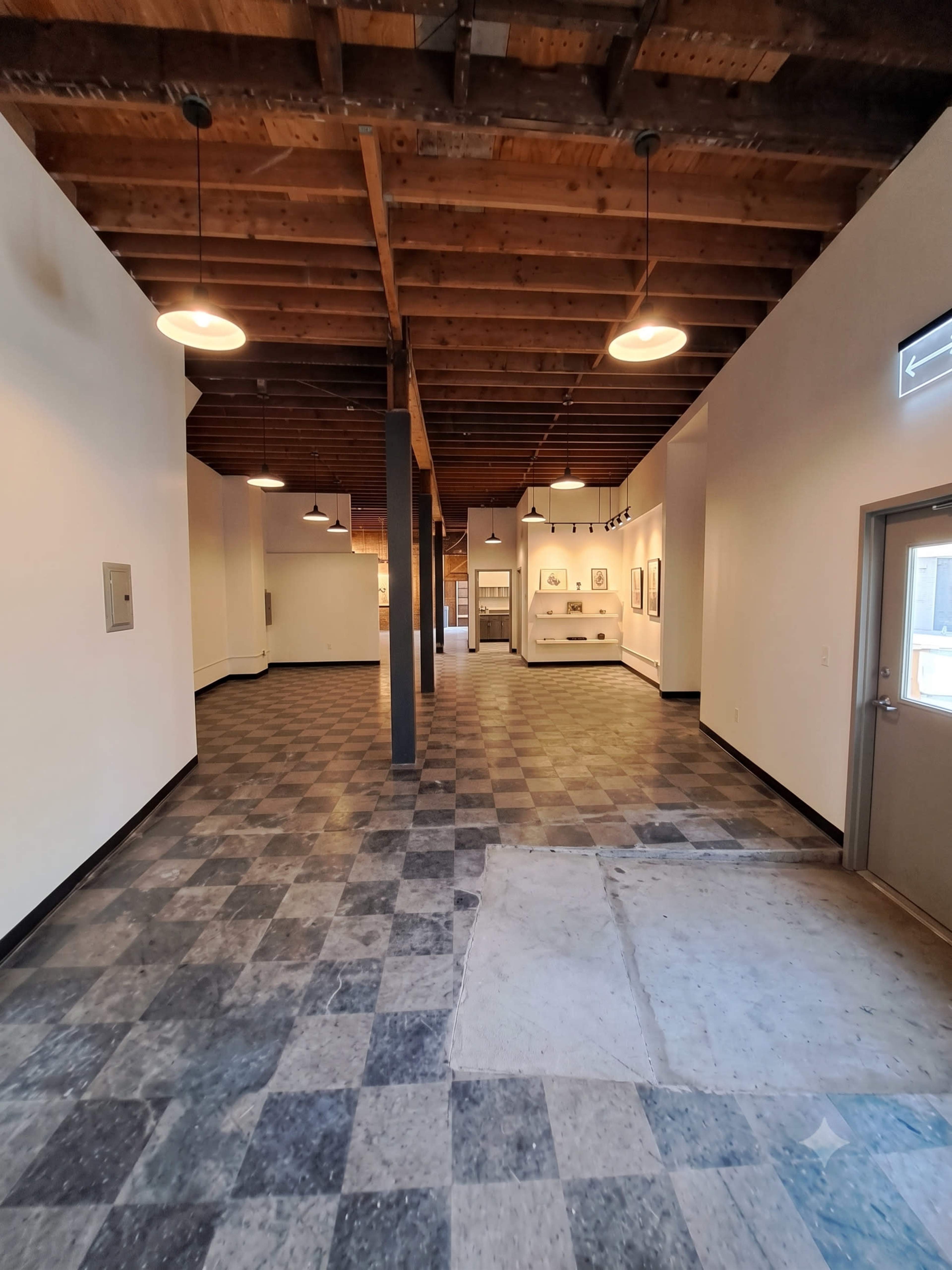 The image shows a spacious, empty interior of a gallery or open room with a checkered floor, exposed wooden beams, and a few wall-mounted light fixtures.