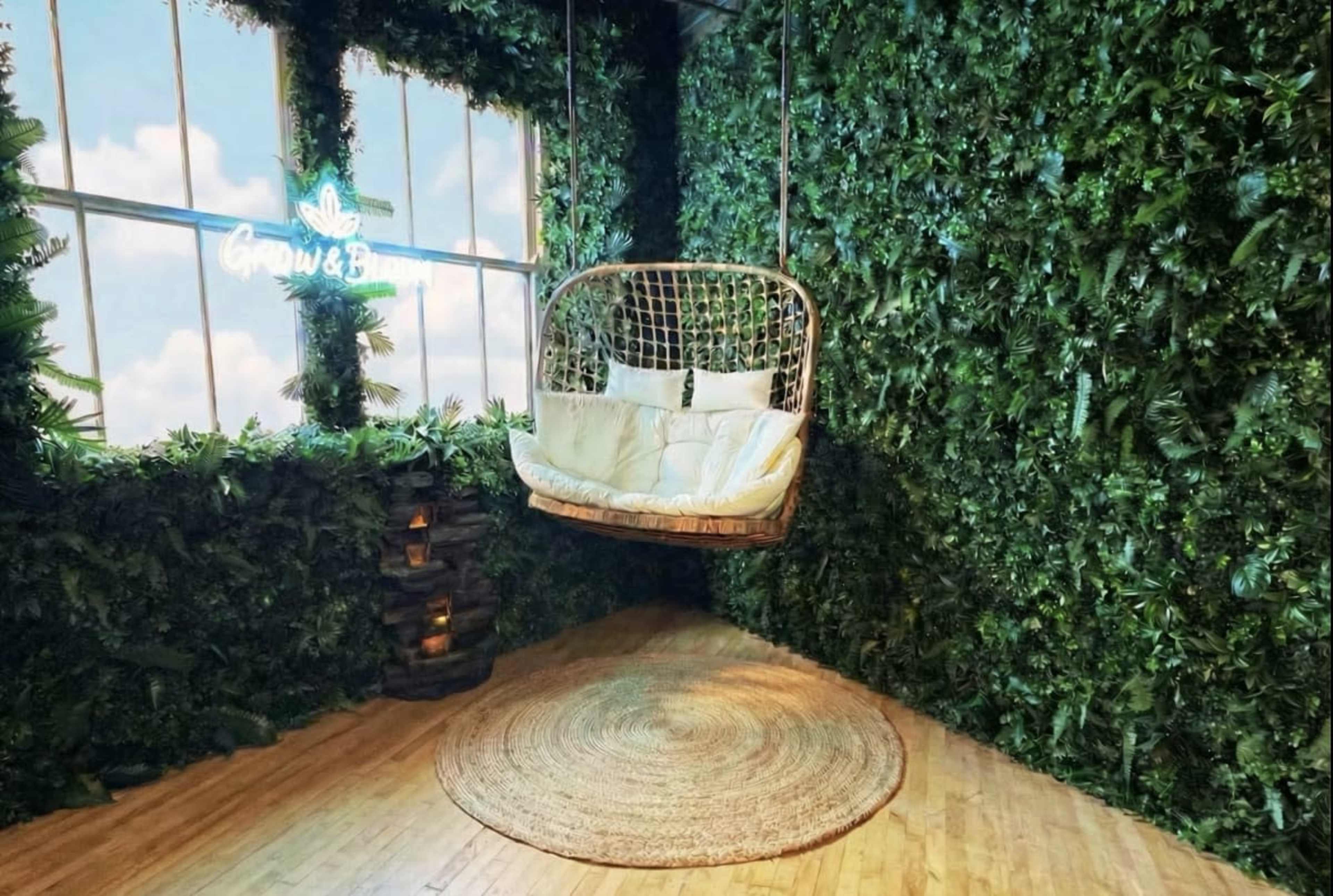 The image shows a cozy swinging chair surrounded by a wall of greenery and a large window with a blue sky backdrop.