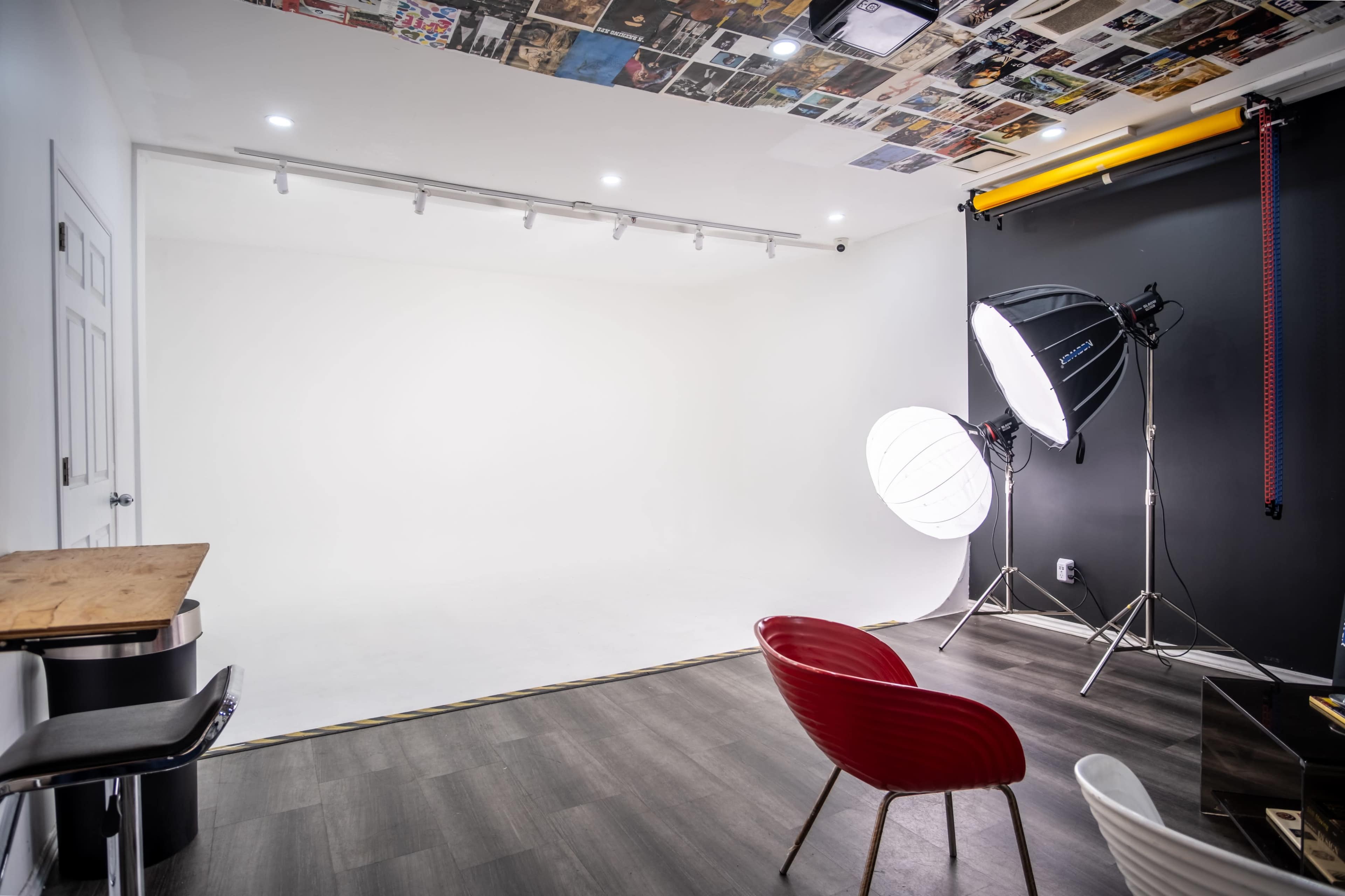 The image shows a photography studio with a blank white backdrop, lighting equipment, and minimalist furniture including a red chair and a bar stool.