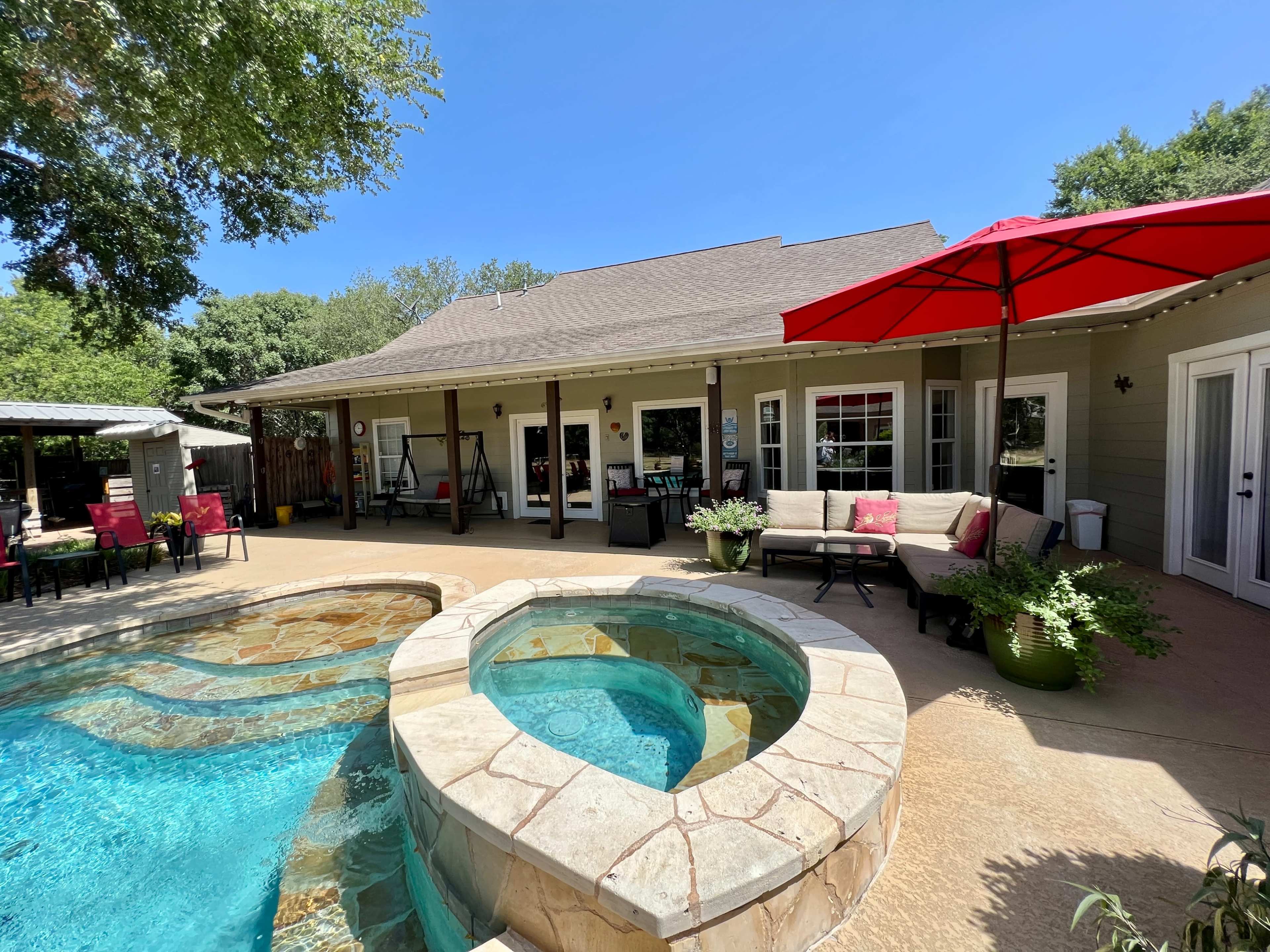 A backyard pool area featuring a hot tub, a shaded seating area with an umbrella, and a house with large windows.