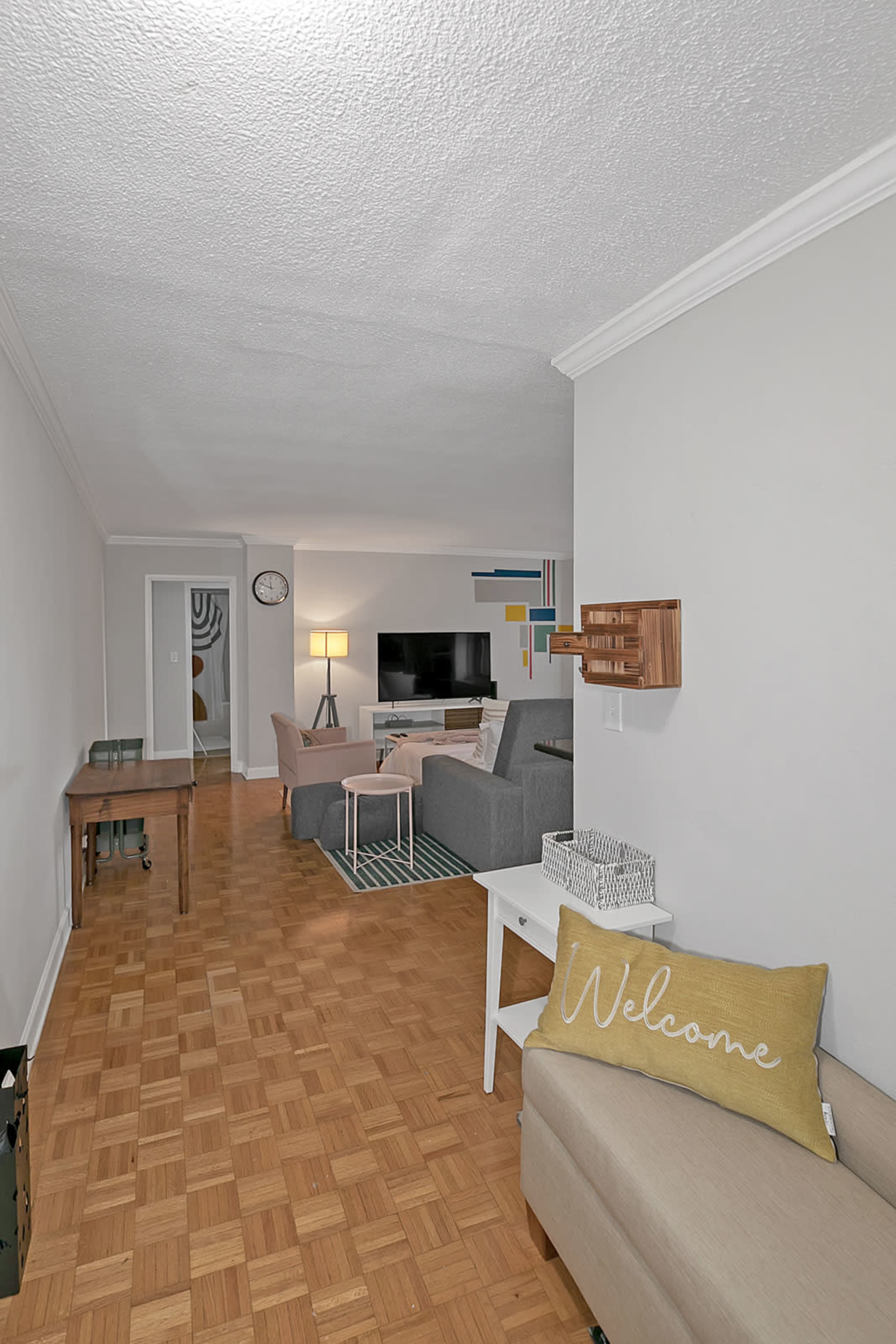 The image shows a living room area featuring a TV, gray sofas, a small white table, and a decorative welcome pillow on a beige couch, with wooden flooring and neutral-colored walls.
