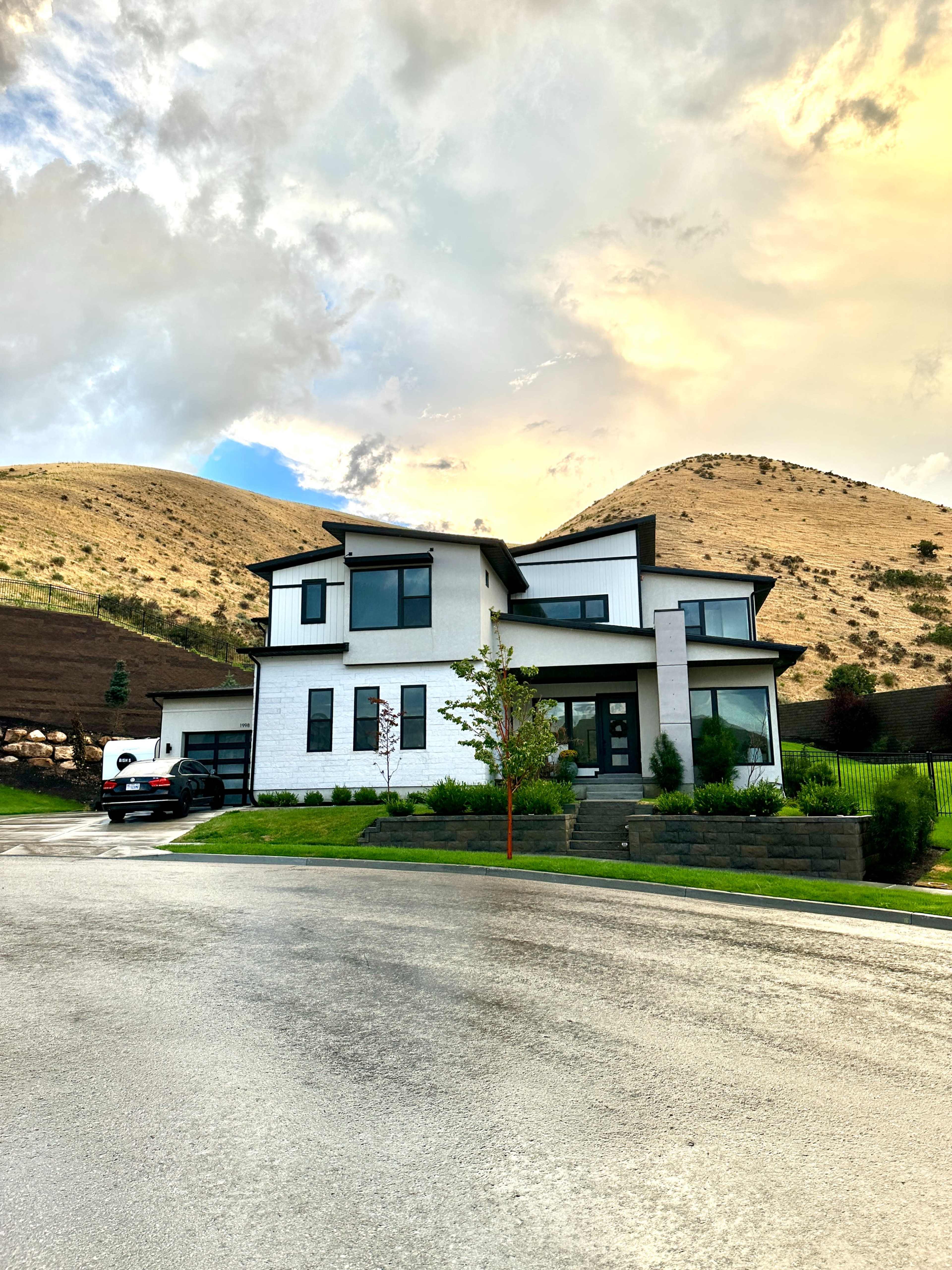 A modern two-story house with a flat roof is situated on a curved road surrounded by rolling hills.