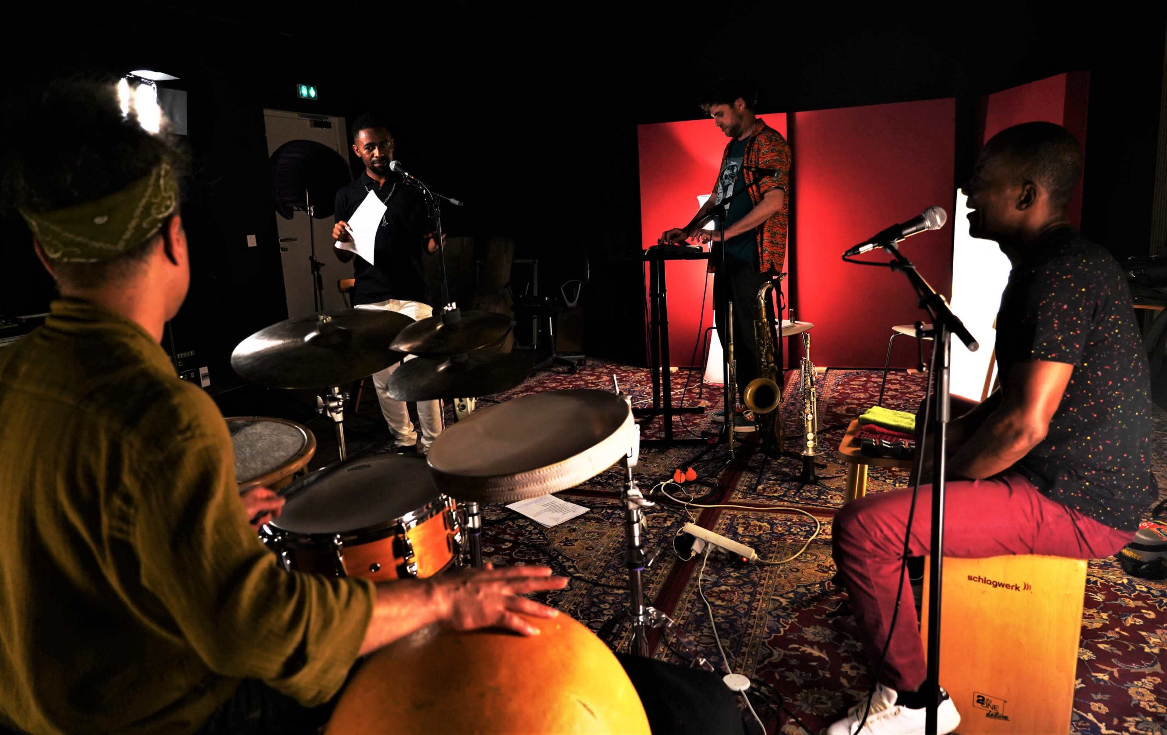 A band performs in a studio, featuring a drummer, a saxophonist, and two musicians at microphones.
