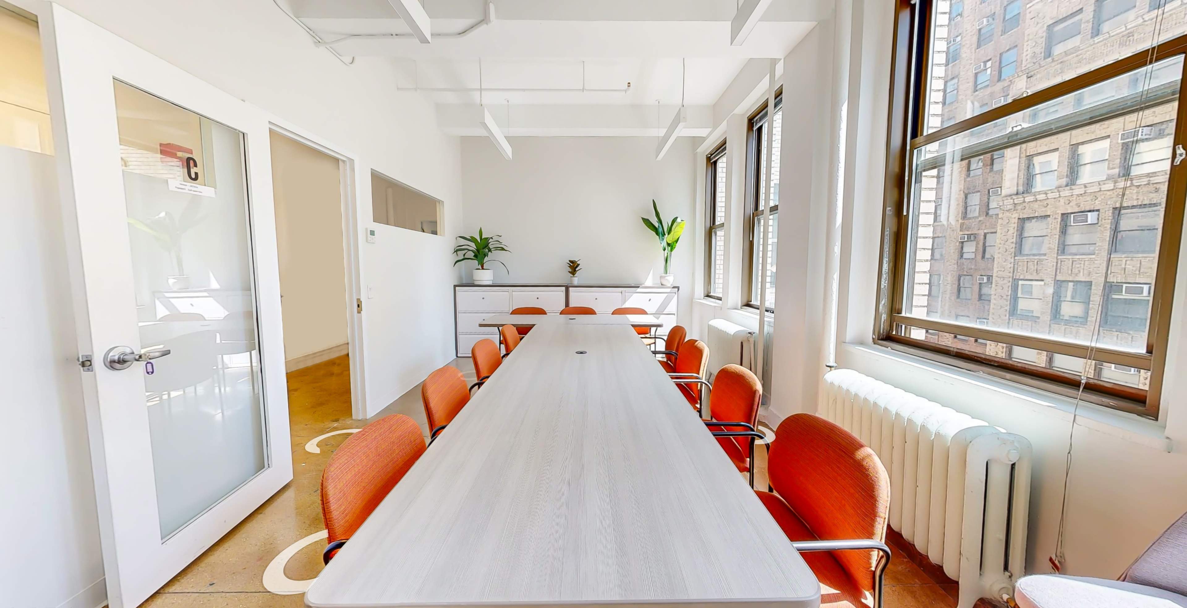 A long conference table with orange chairs is situated in a bright, modern meeting room with large windows and a potted plant.