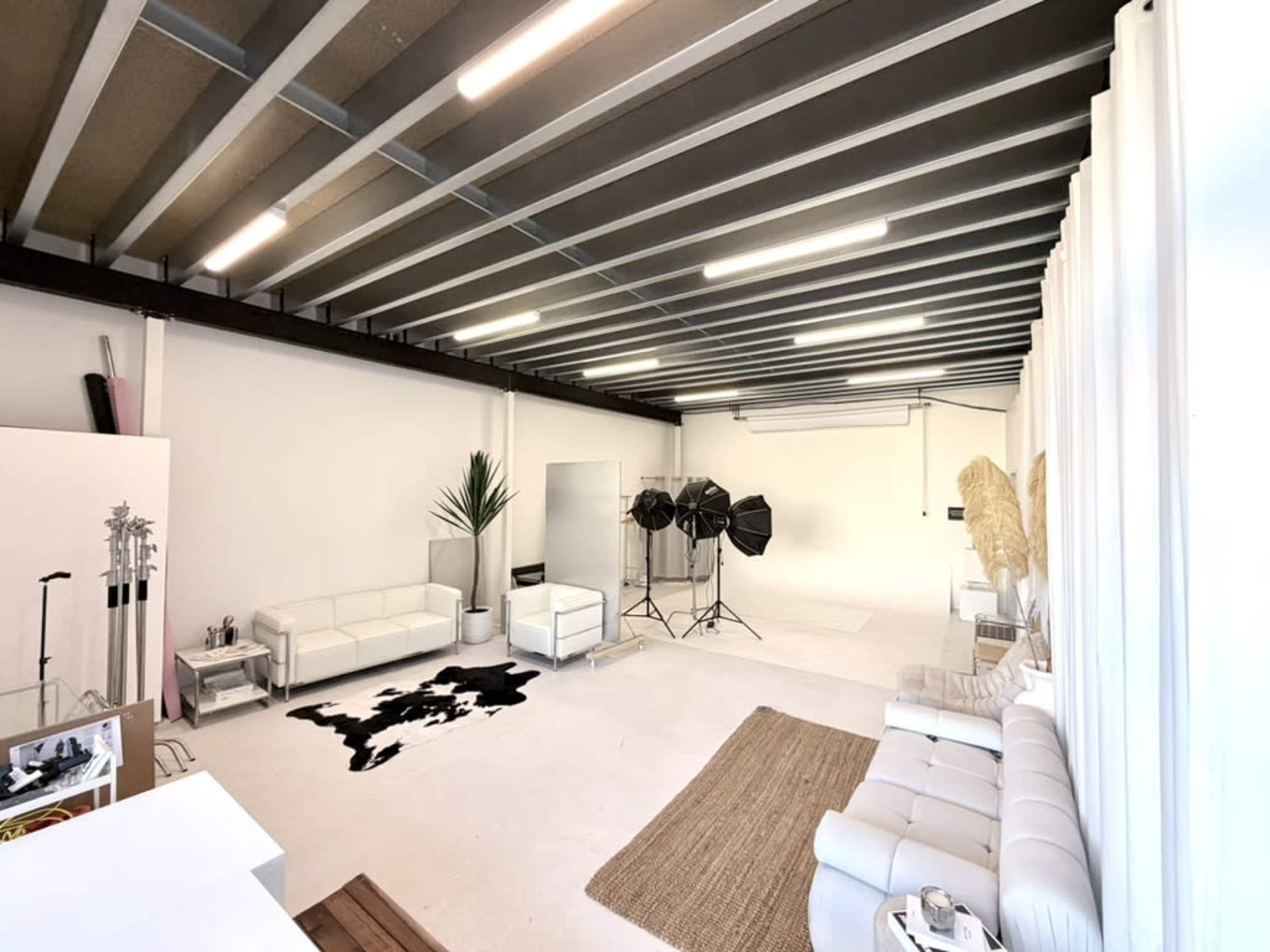 The image shows a spacious, well-lit studio with white walls, modern furniture, and photography equipment set up in one corner.