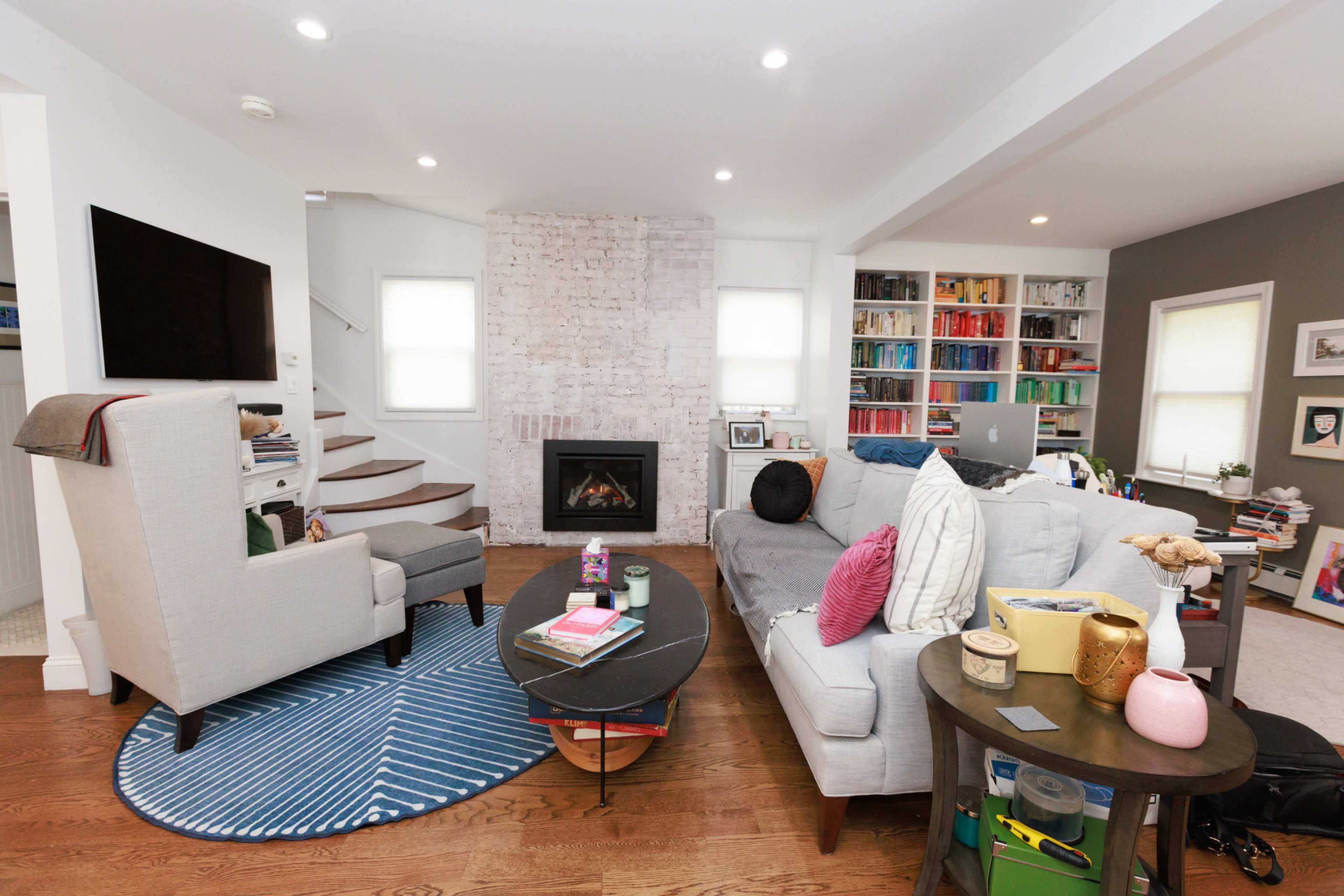 A modern living room features a gray sofa, a cozy fireplace, a coffee table, shelves filled with books, and a staircase leading to another level.