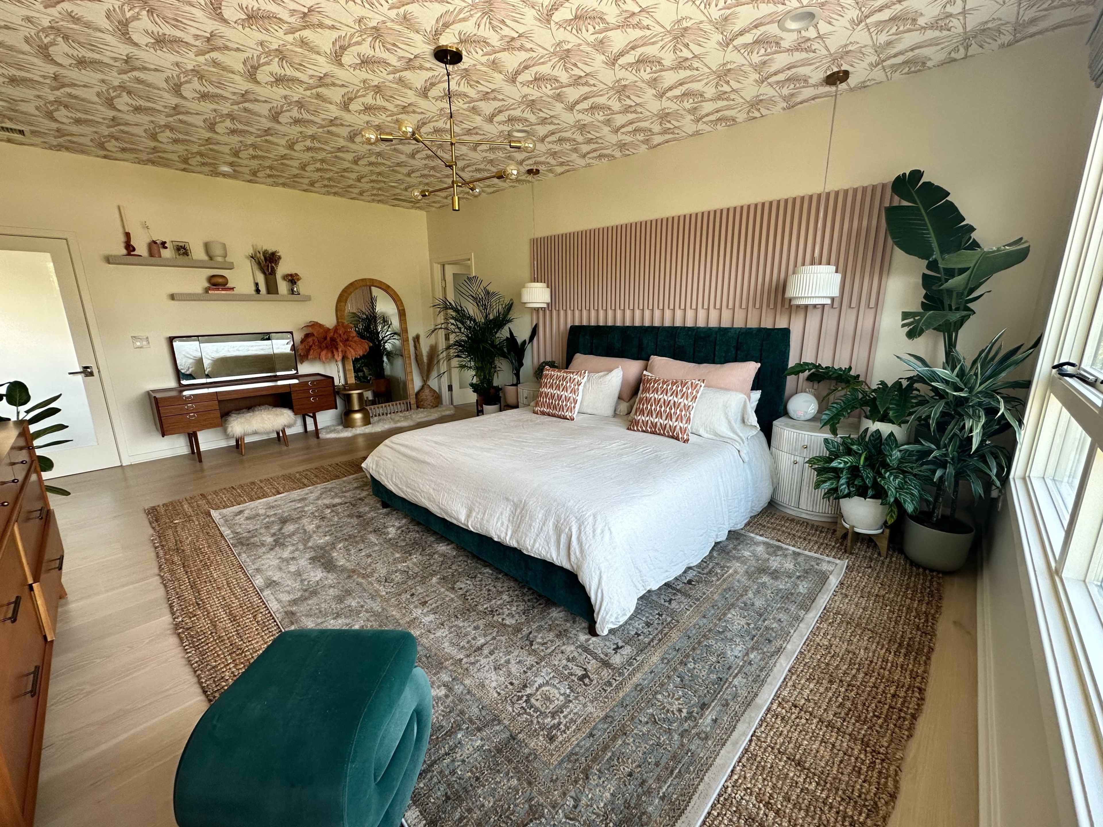 A spacious bedroom featuring a large upholstered bed, decorative pillows, various plants, a patterned ceiling, and a combination of modern furniture pieces.