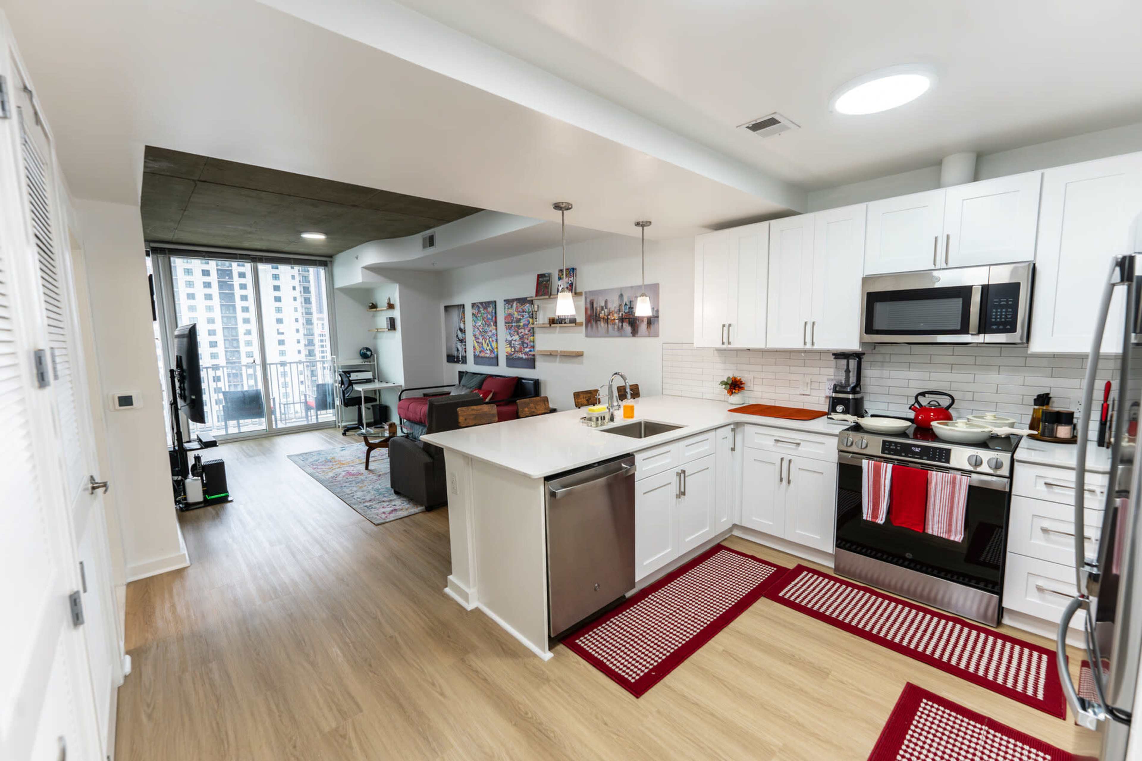 The image shows a modern kitchen with white cabinetry and a stainless steel fridge, adjacent to a living area featuring a sofa and a balcony.