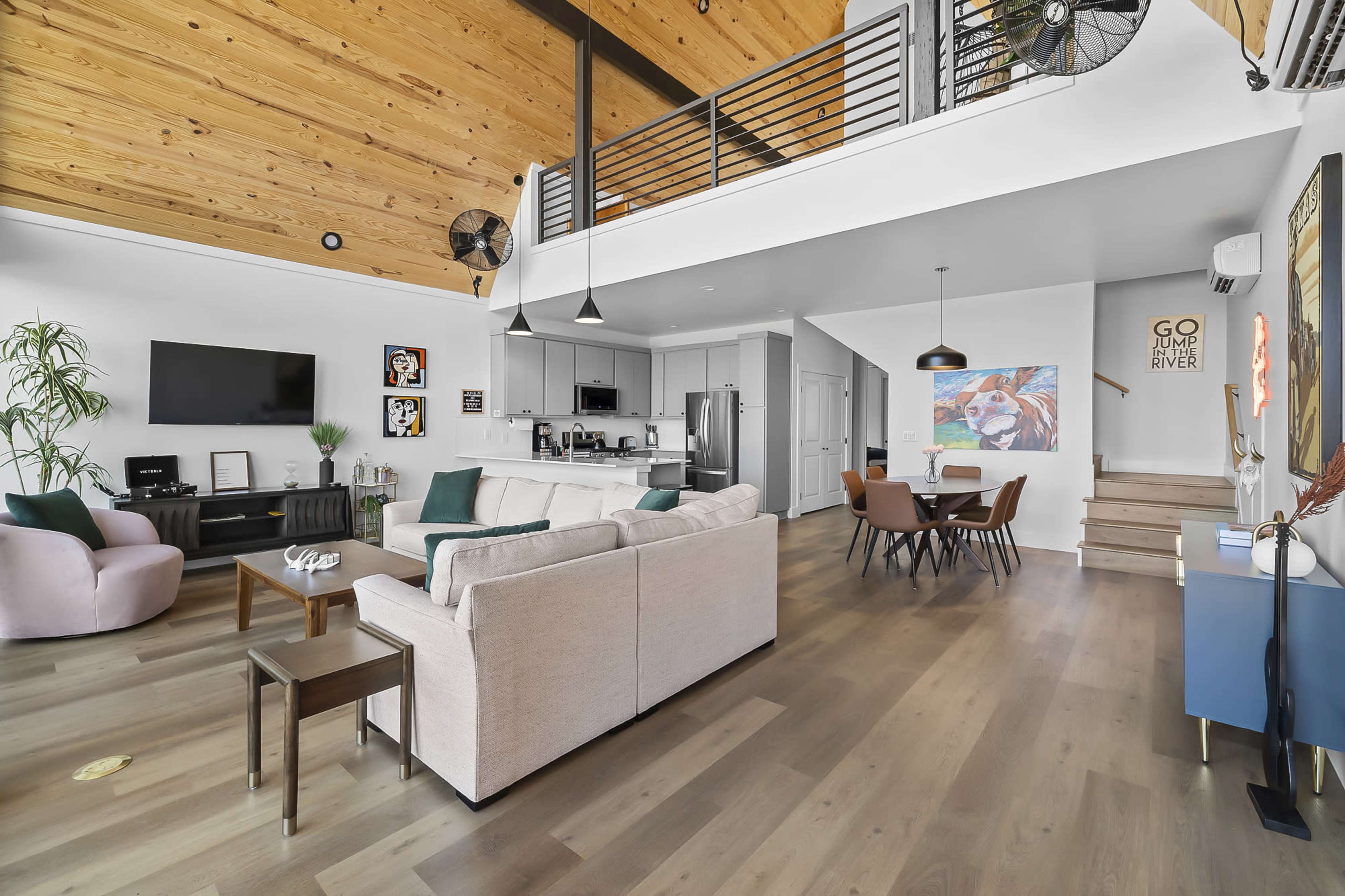 A modern open-concept living space features a well-furnished lounge area, dining table, and kitchen under a high wooden ceiling.
