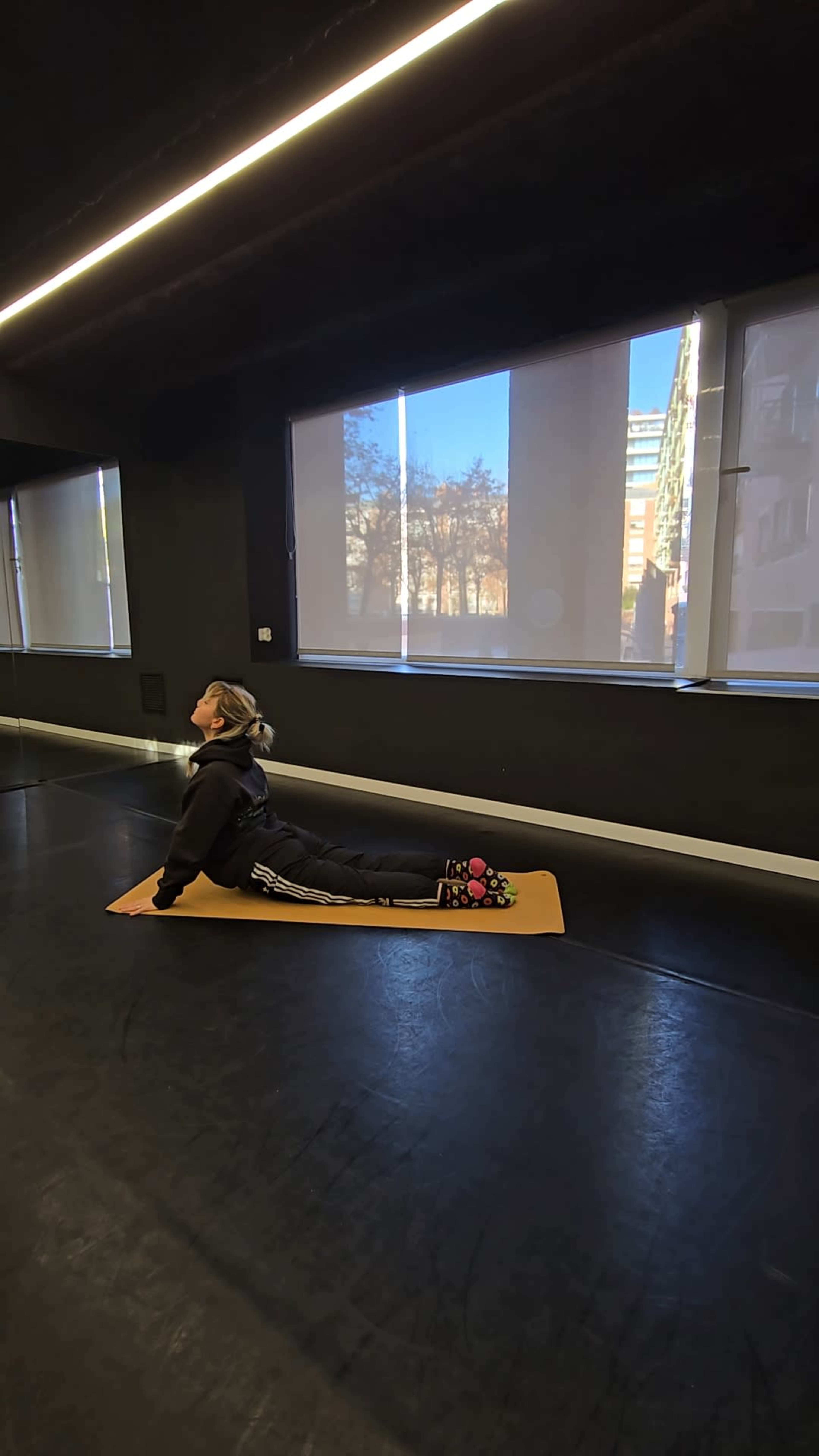 A person in athletic wear performs a yoga pose on a mat in a studio with large windows.