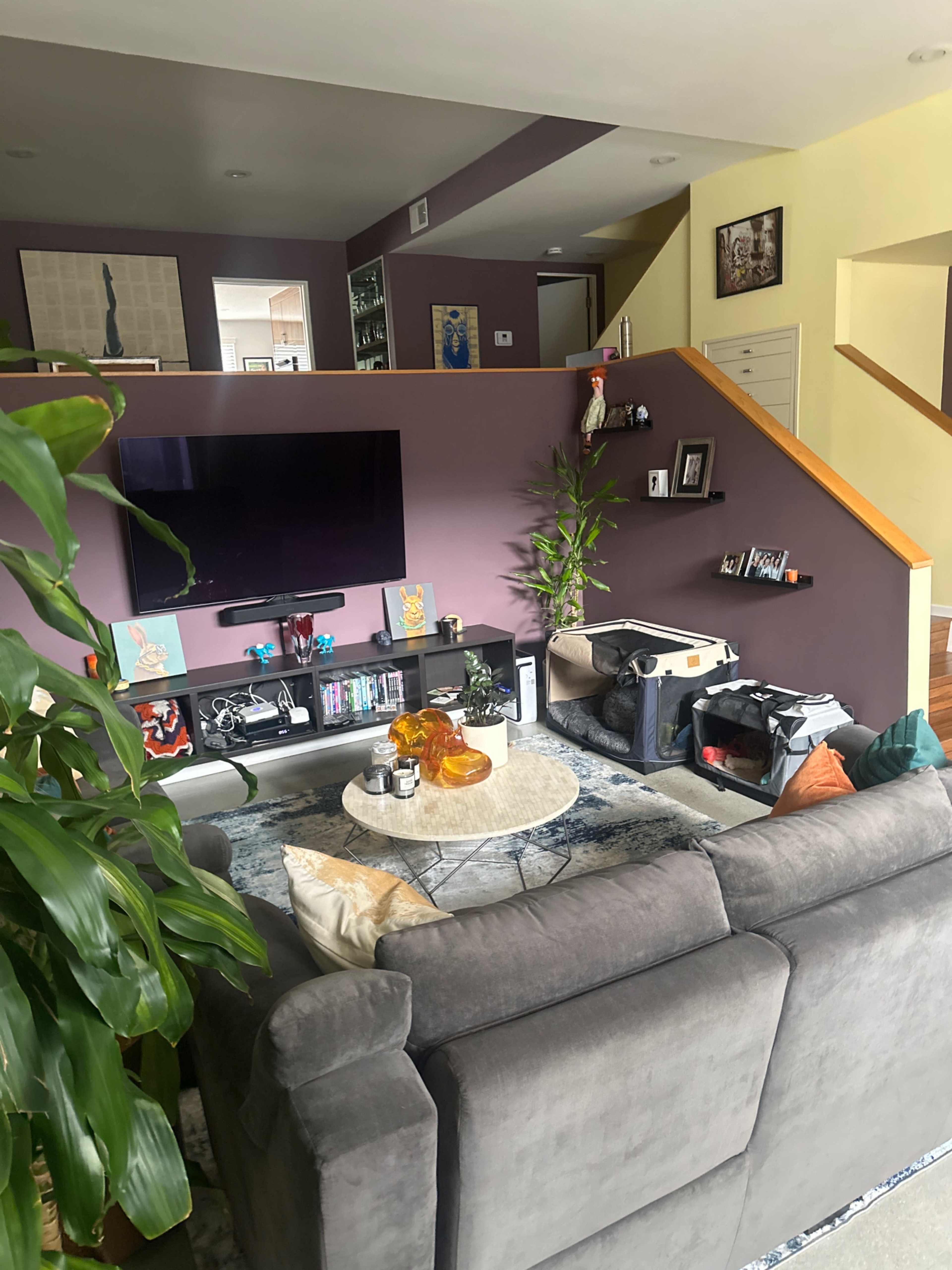 A living room features a gray sofa, a glass coffee table, a large television on a wall, and stairs leading to an upper level.